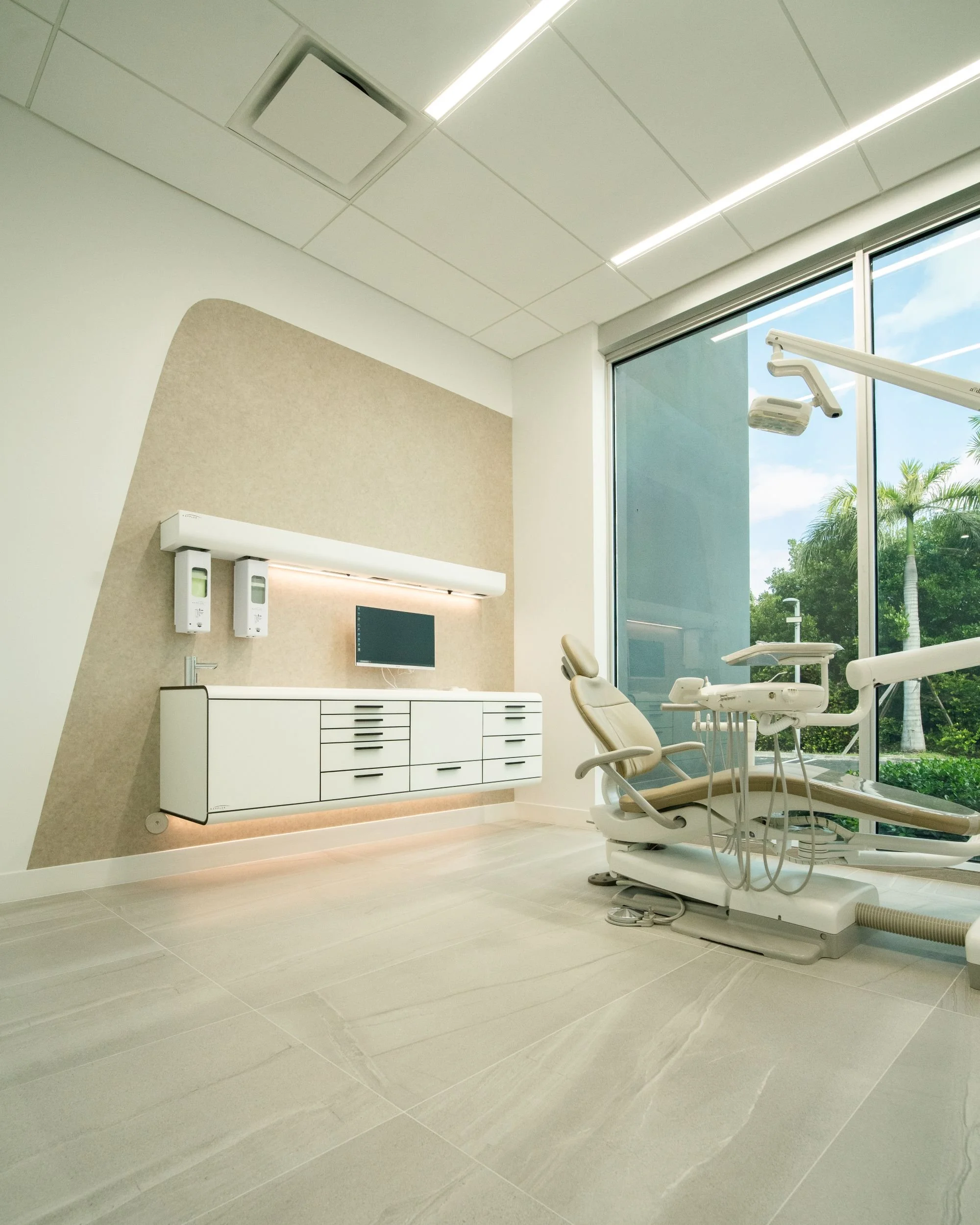 A dental examination room with a dental chair, large window showing palm trees outside, and medical equipment on the wall.