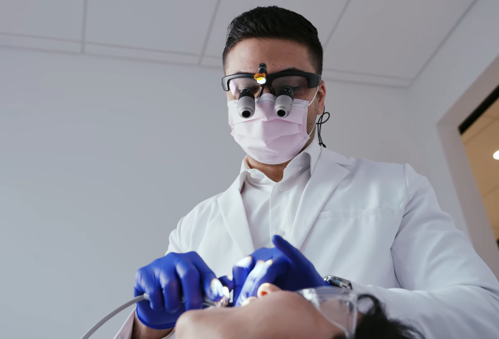 Dentist wearing a face mask and magnifying loupes examines a patient's teeth with dental instruments.