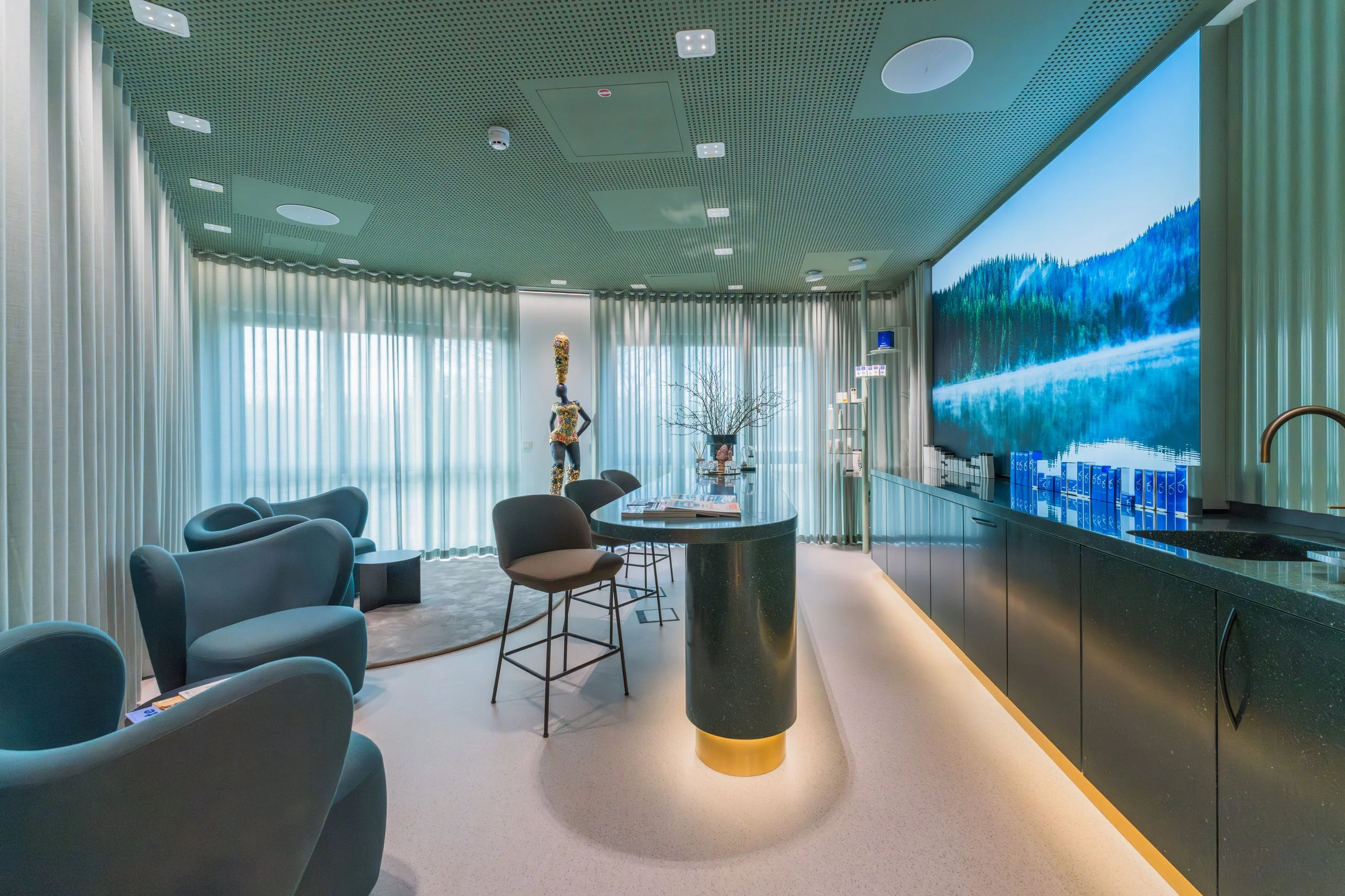 Modern living room with a large digital screen showing a lake landscape, a round black table, chairs, and a sculpture of a woman with a tall headpiece, next to a window with sheer curtains and a decorative tree.