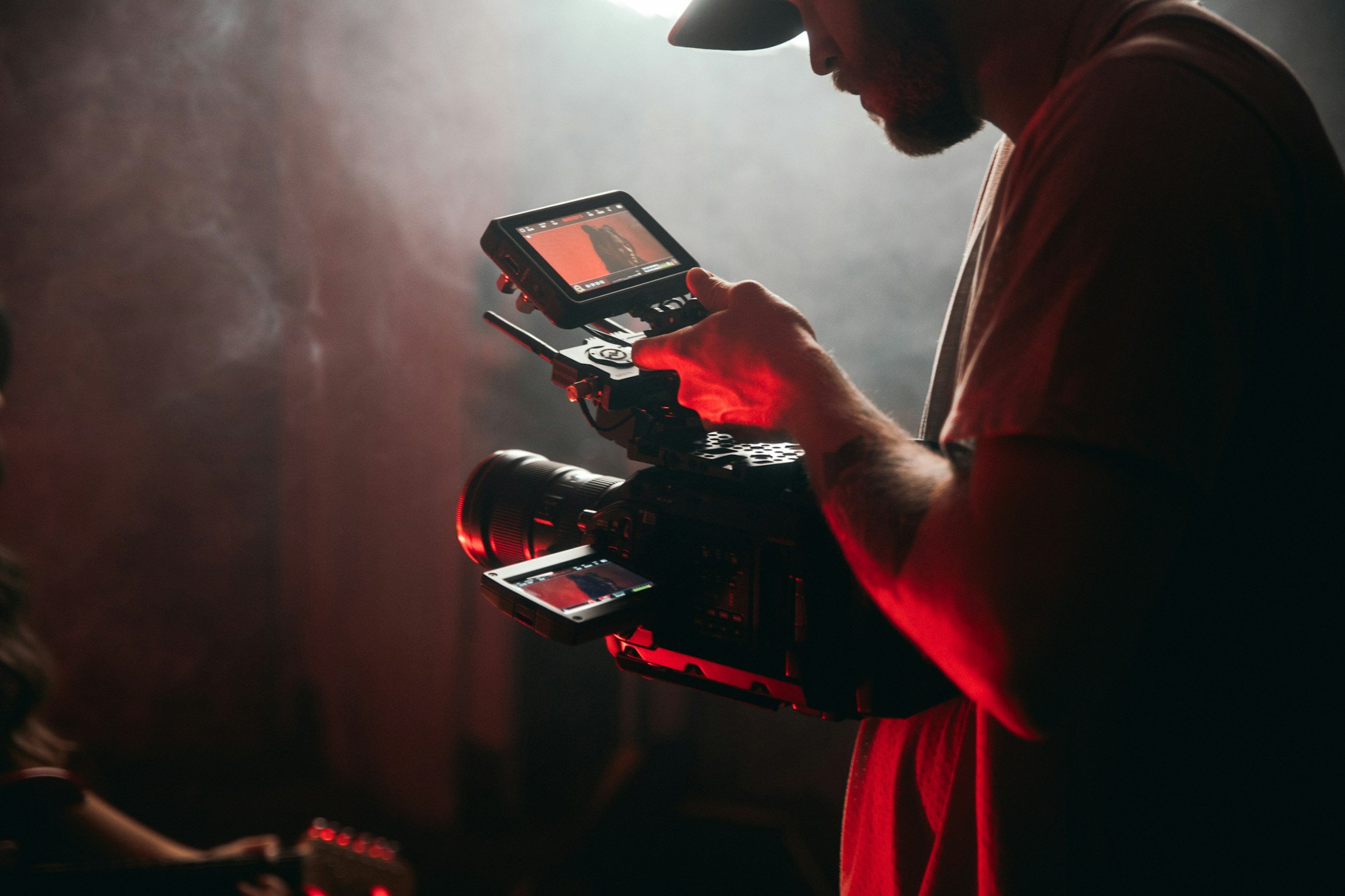 A man in a hat is operating a professional video camera in a dimly lit environment. The camera has a mounted monitor and an external light, with red ambient lighting.