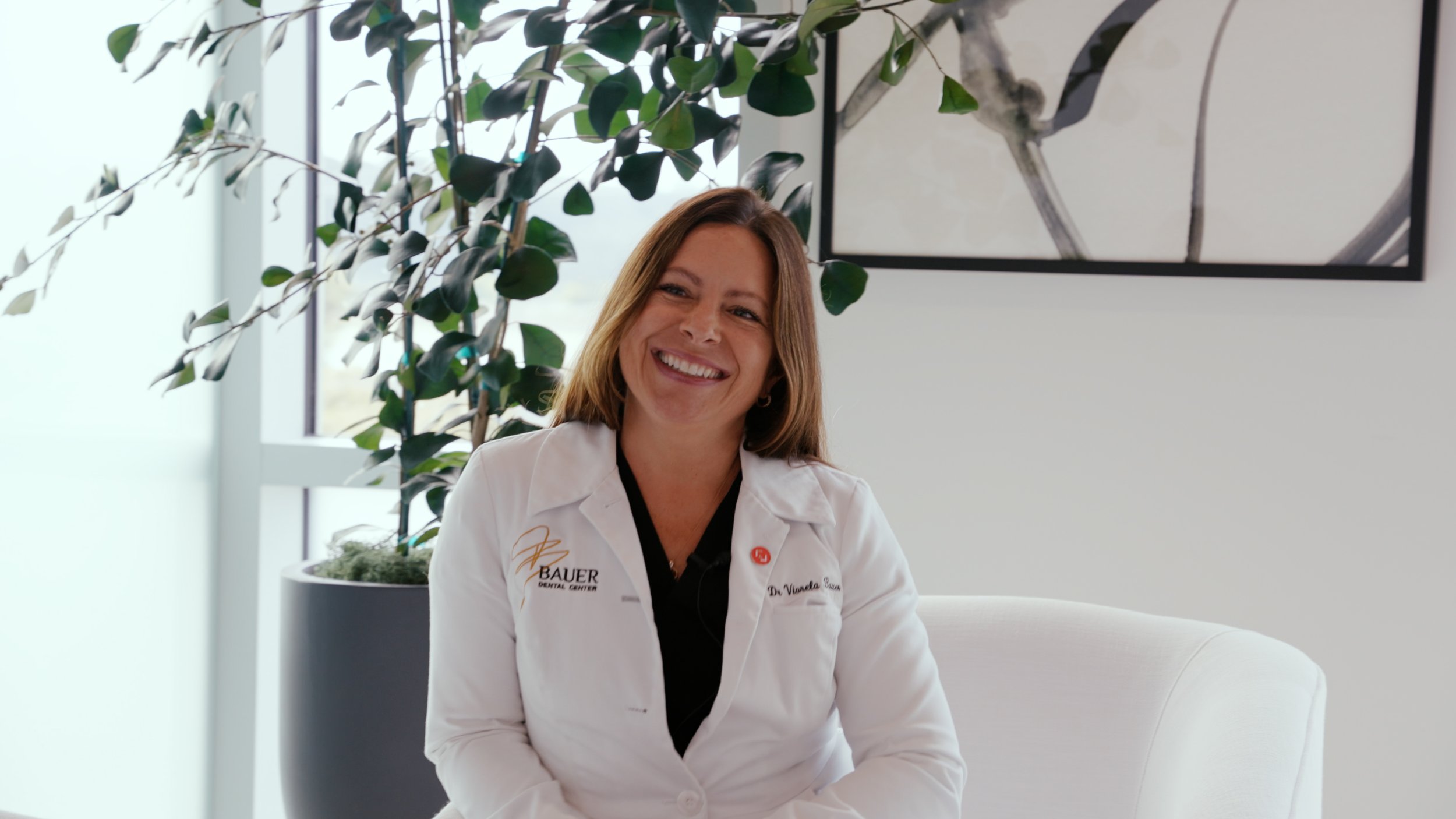 A smiling woman in a white lab coat sitting on a white chair near a window with a large indoor plant behind her.