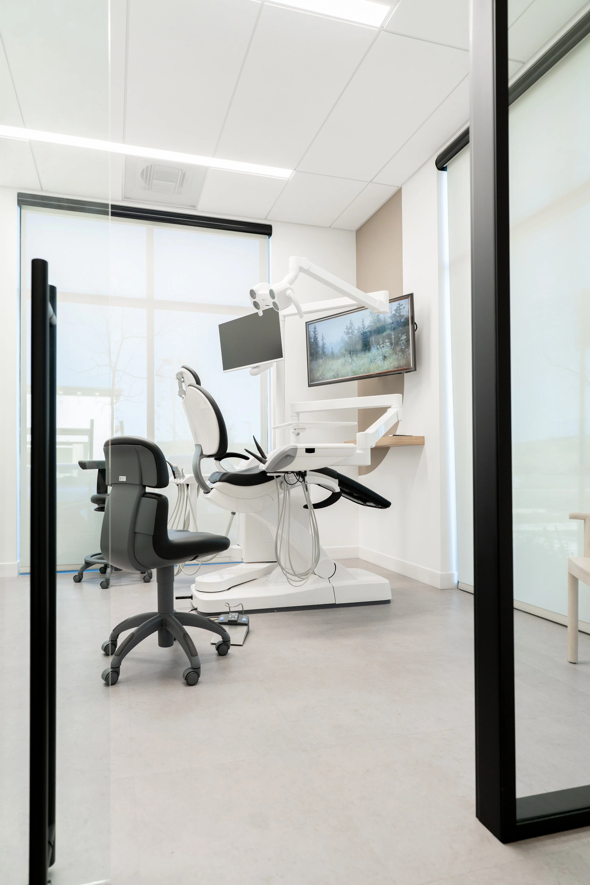 A medical examination room with a dental chair, overhead dental light, monitor, and large window showing trees outside.