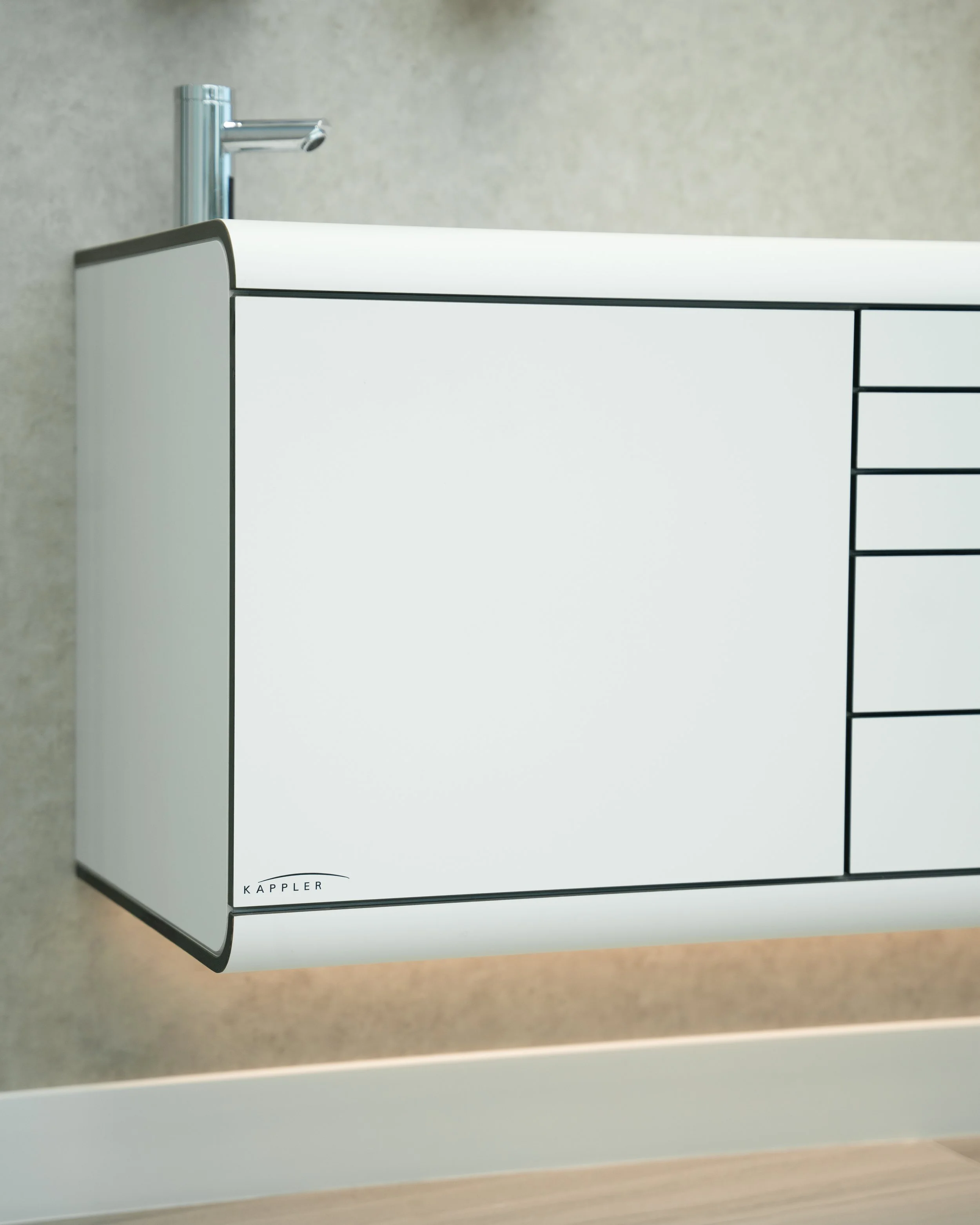 Close-up of a modern white cabinet with black accents labeled 'Kappler' at the bottom, featuring a silver faucet on top, set against a gray wall.