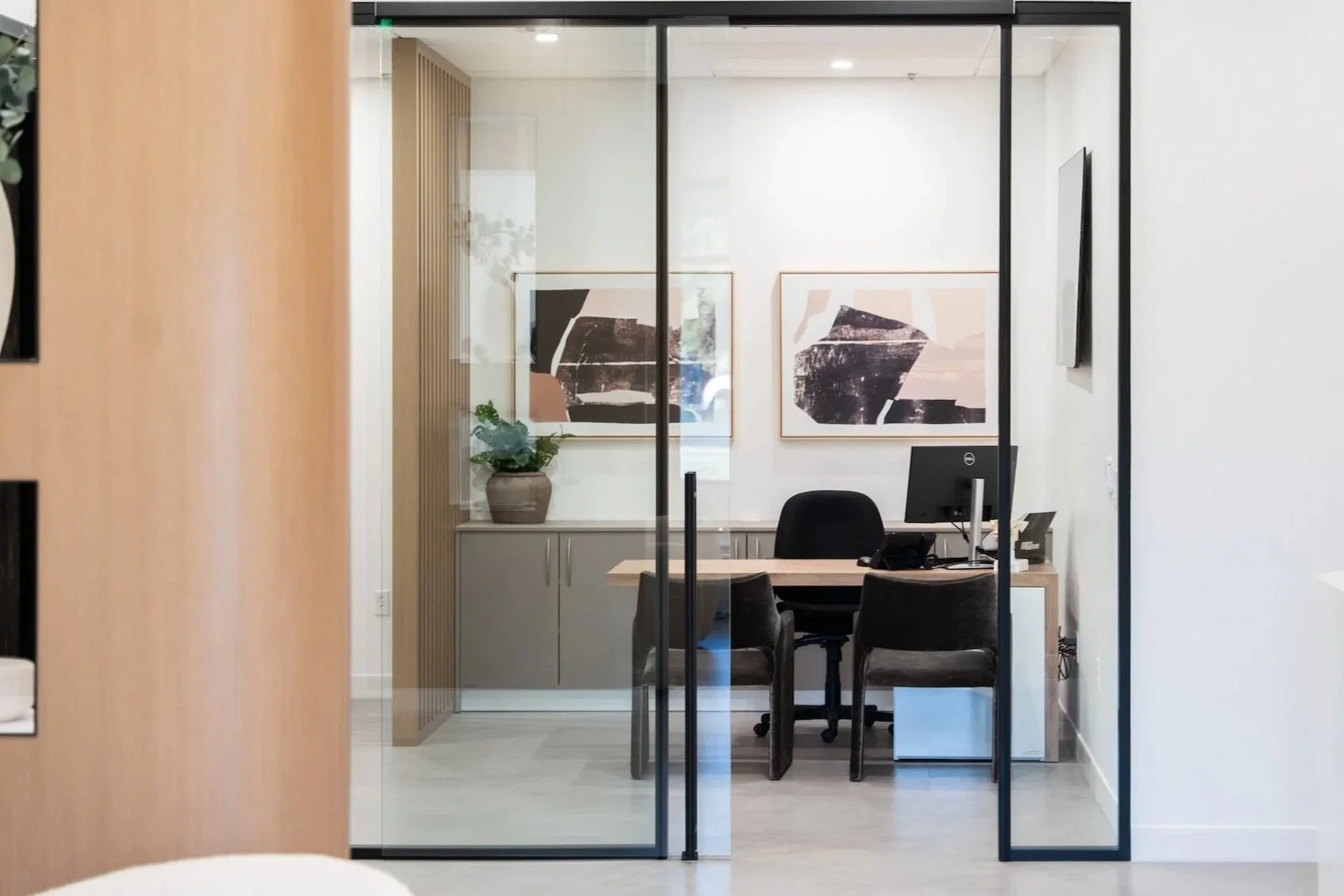 An office with glass walls, a wooden desk, three black chairs, a computer, a telephone, against a white wall decorated with abstract artwork, and a plant in a gray pot on a gray cabinet.