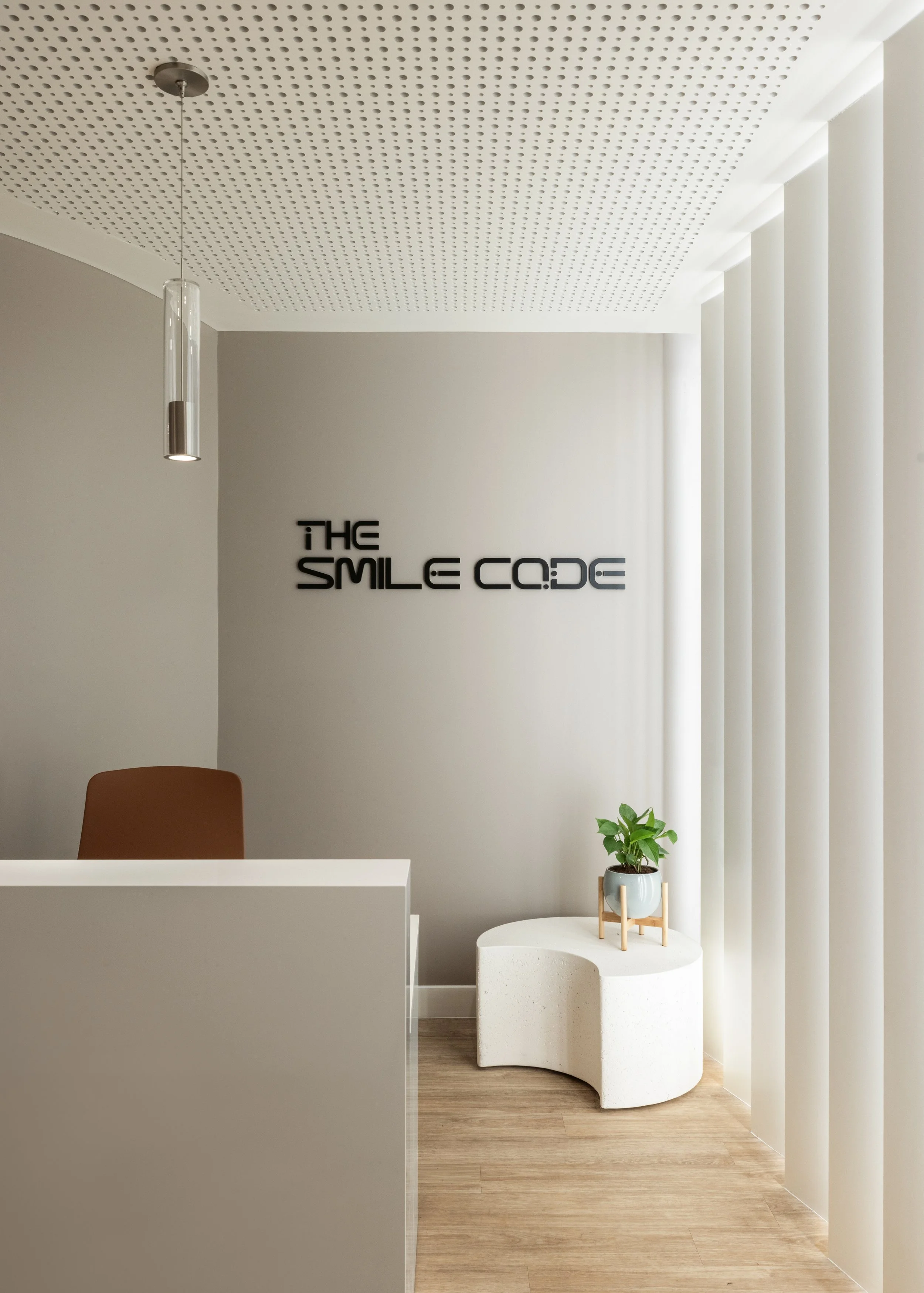 Modern reception area with white walls, wooden flooring, a brown chair, a potted green plant on a white side table, and a decorative wall with vertical paneling. A hanging pendant light is visible near the ceiling, which has perforated acoustic panels. The wall displays the text 'THE SMILE CODE' in black lettering.
