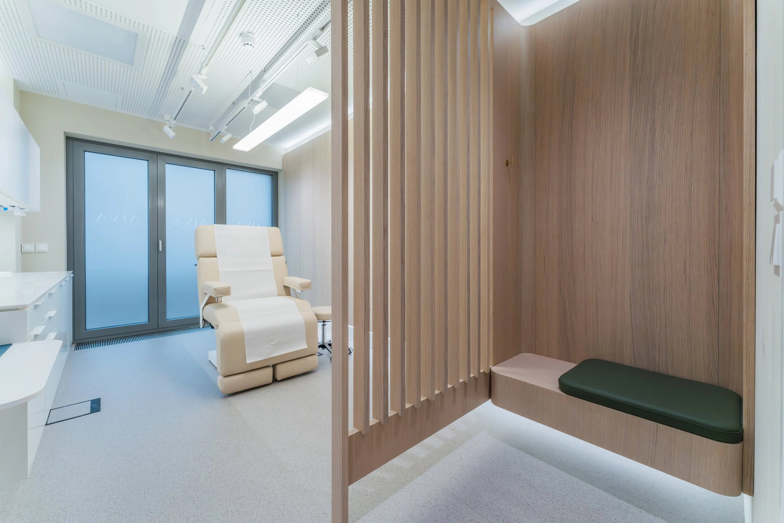 A modern medical or consultation room with a reclining chair covered with a paper sheet, a wooden privacy partition with a cushioned bench, and large frosted glass windows in the background.