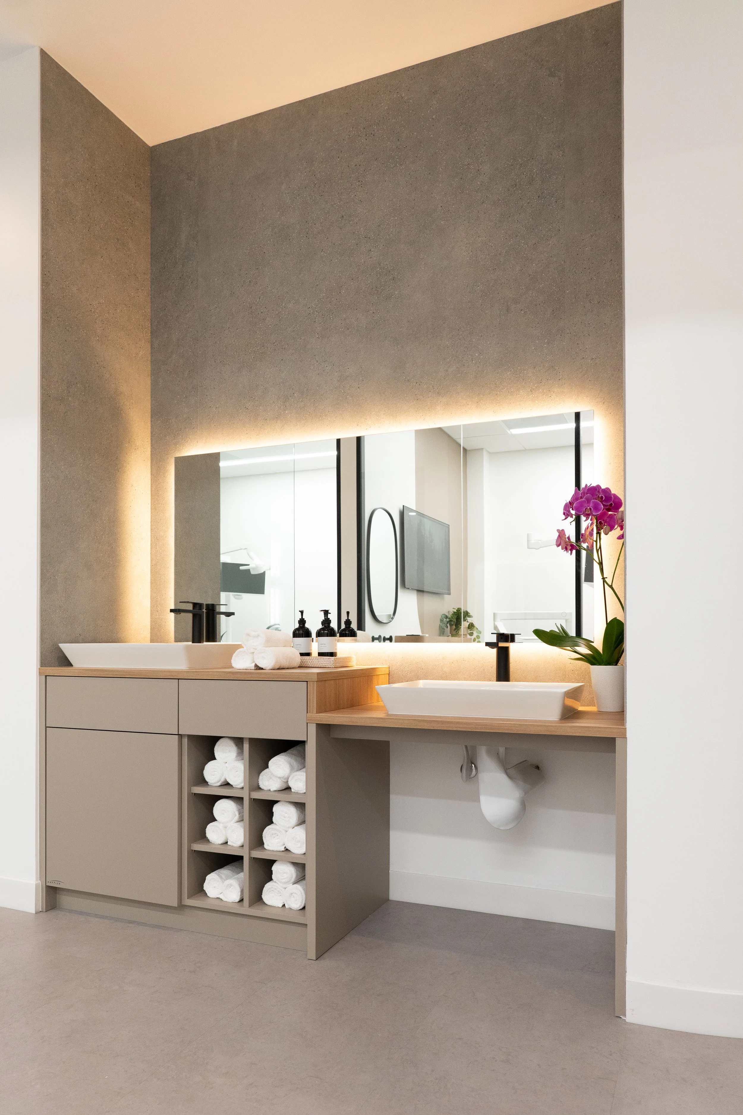 Modern bathroom with beige and gray color scheme, featuring a large mirror, a double sink vanity with black faucets, and a shelf with rolled white towels. A potted orchid is placed on the right side of the vanity. The gray wall has accent lighting, a