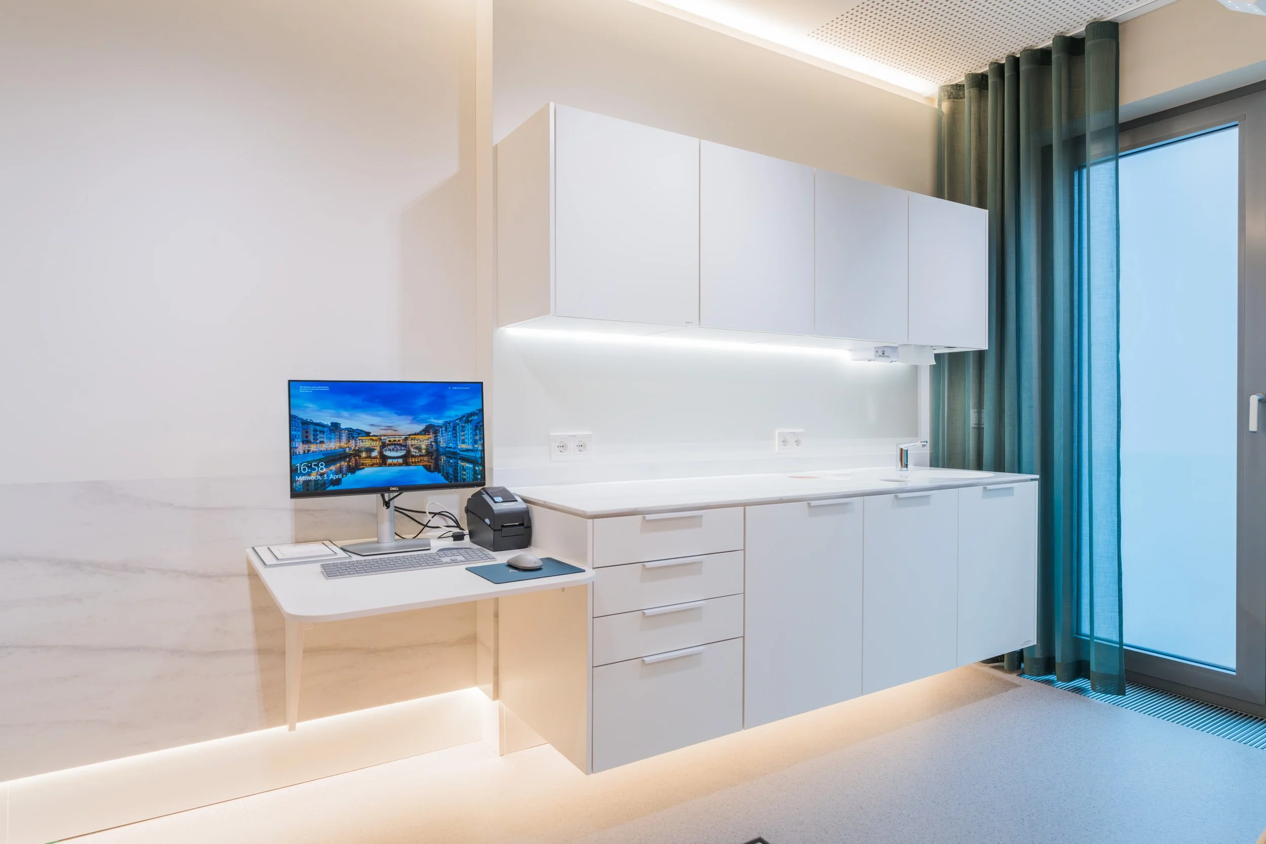 Modern kitchen with white cabinets, a small desk with a computer monitor, keyboard, mouse, and a printer, and a sliding glass door with blue curtains.