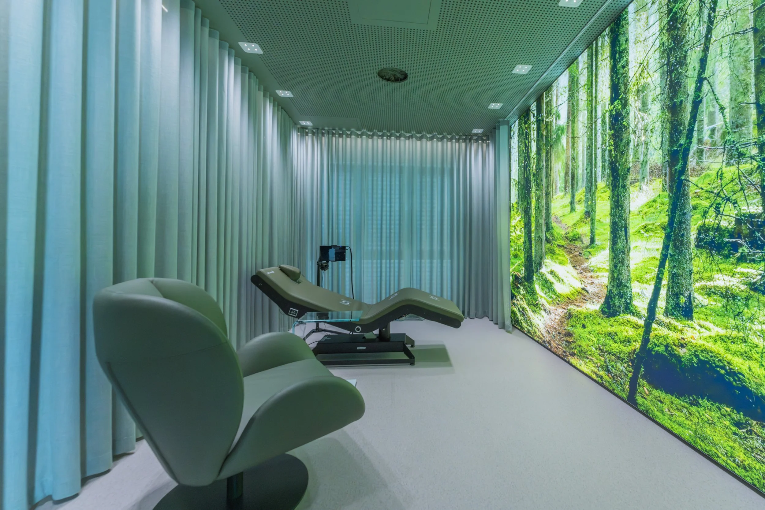 Medical room with two chairs and a treatment bed facing a large wall screen displaying a lush green forest scene