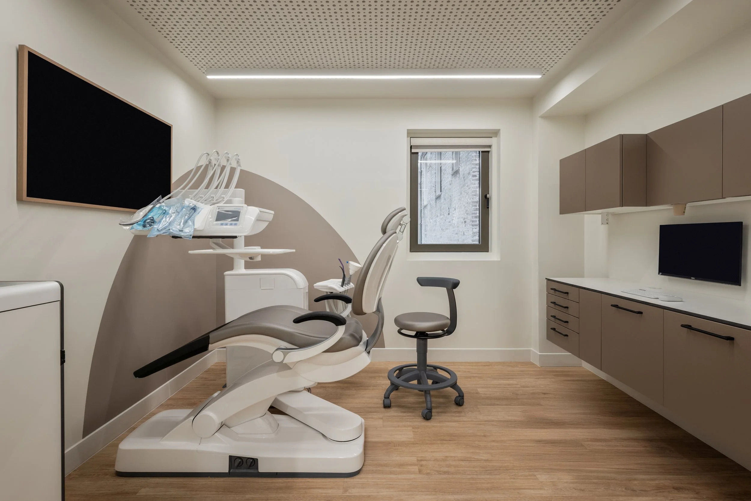 Dental clinic room with dental chair, dental tools, cabinets, a wall-mounted monitor, window, and clean, modern decor.