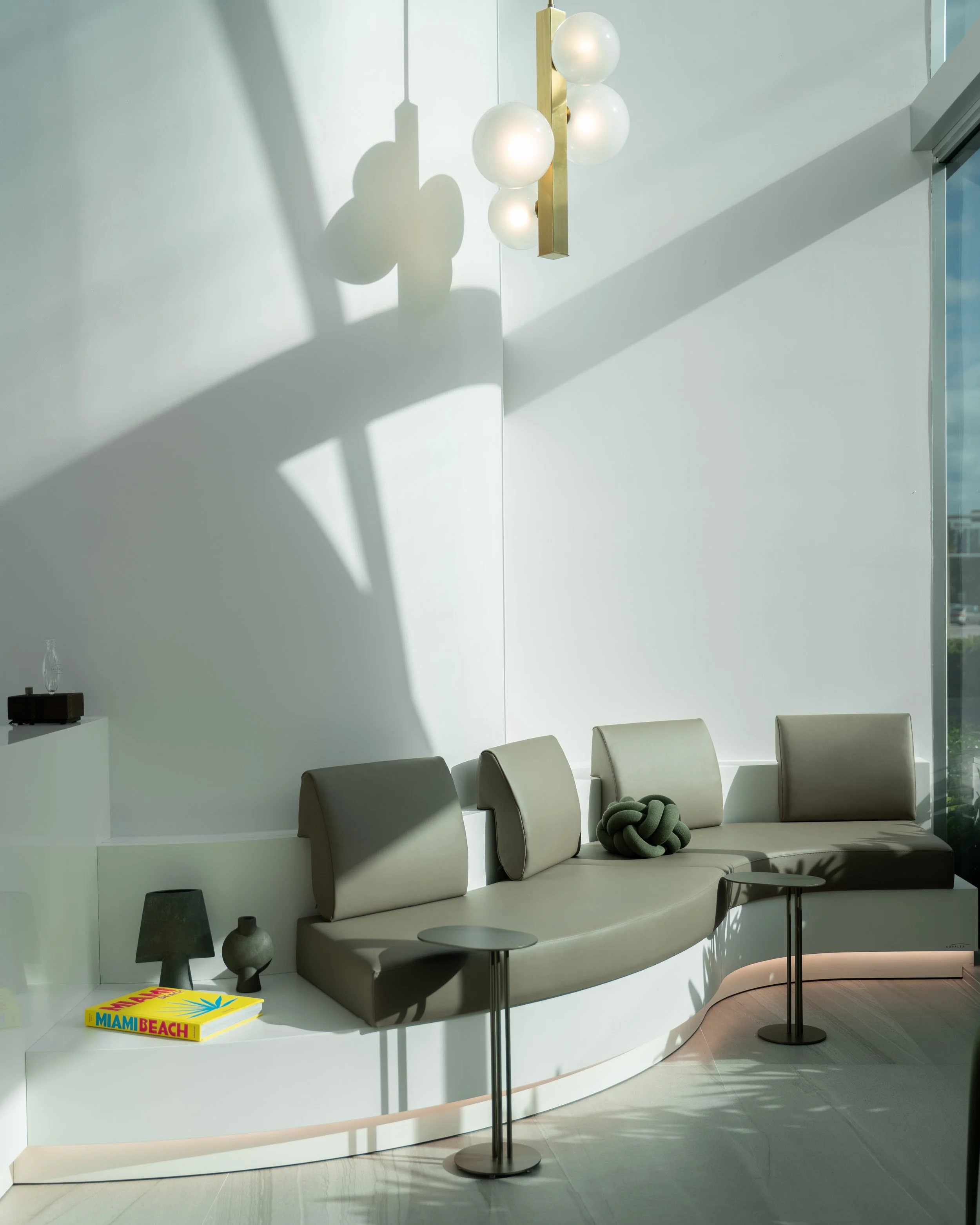 Modern interior with white wall, ceiling lamp casting shadows, beige sofa with three sections, a black vase, a yellow Miami Beach book, black vases, and small round tables.