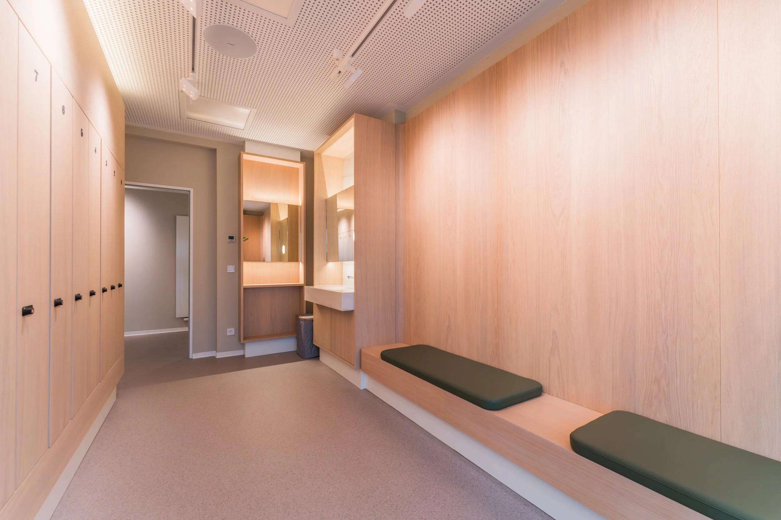 A clean, modern locker room with wooden lockers on the left, a wooden bench with green cushions on the right, and a small vanity area with mirrors and a sink in the background.