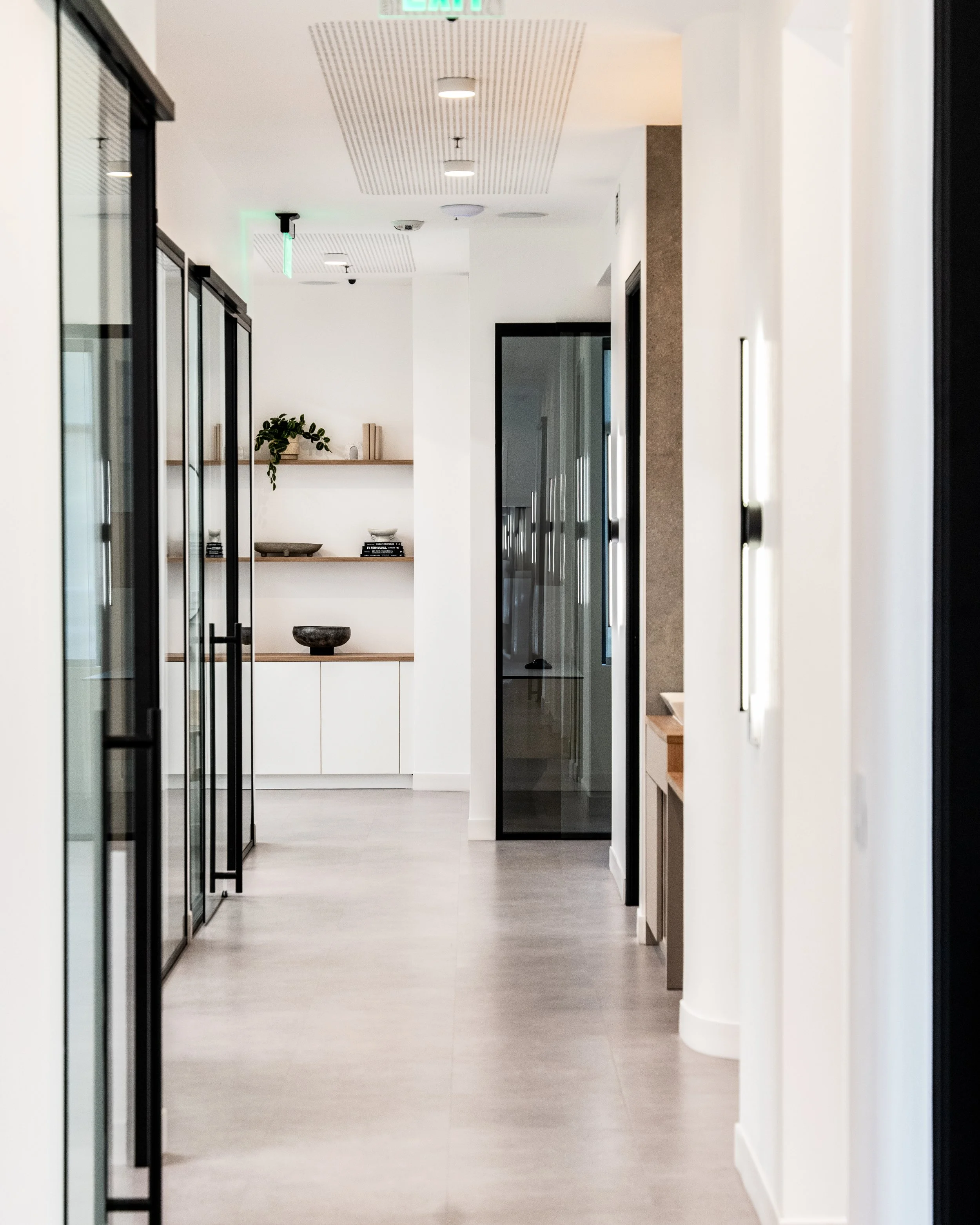 A modern, minimalist hallway in a building with glass doors on either side, white walls, and light-colored wood or laminate flooring. There is a shelf with decorative items and plants on the left side, and the ceiling has recessed lighting and a green exit sign.