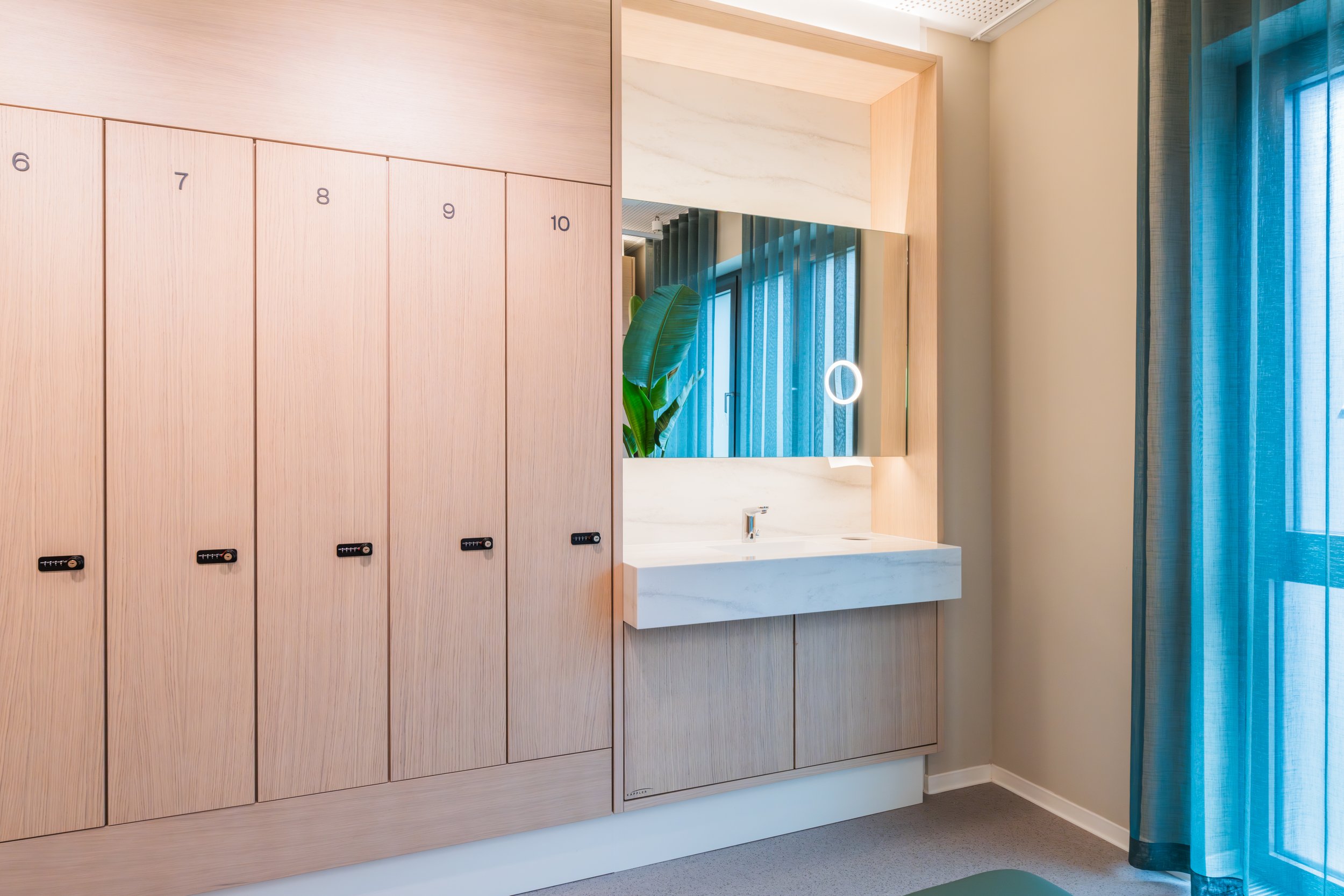 Bathroom with wooden lockers numbered 6 to 10, a mirror, a sink with a white marble countertop, blue curtains, and a large green plant.