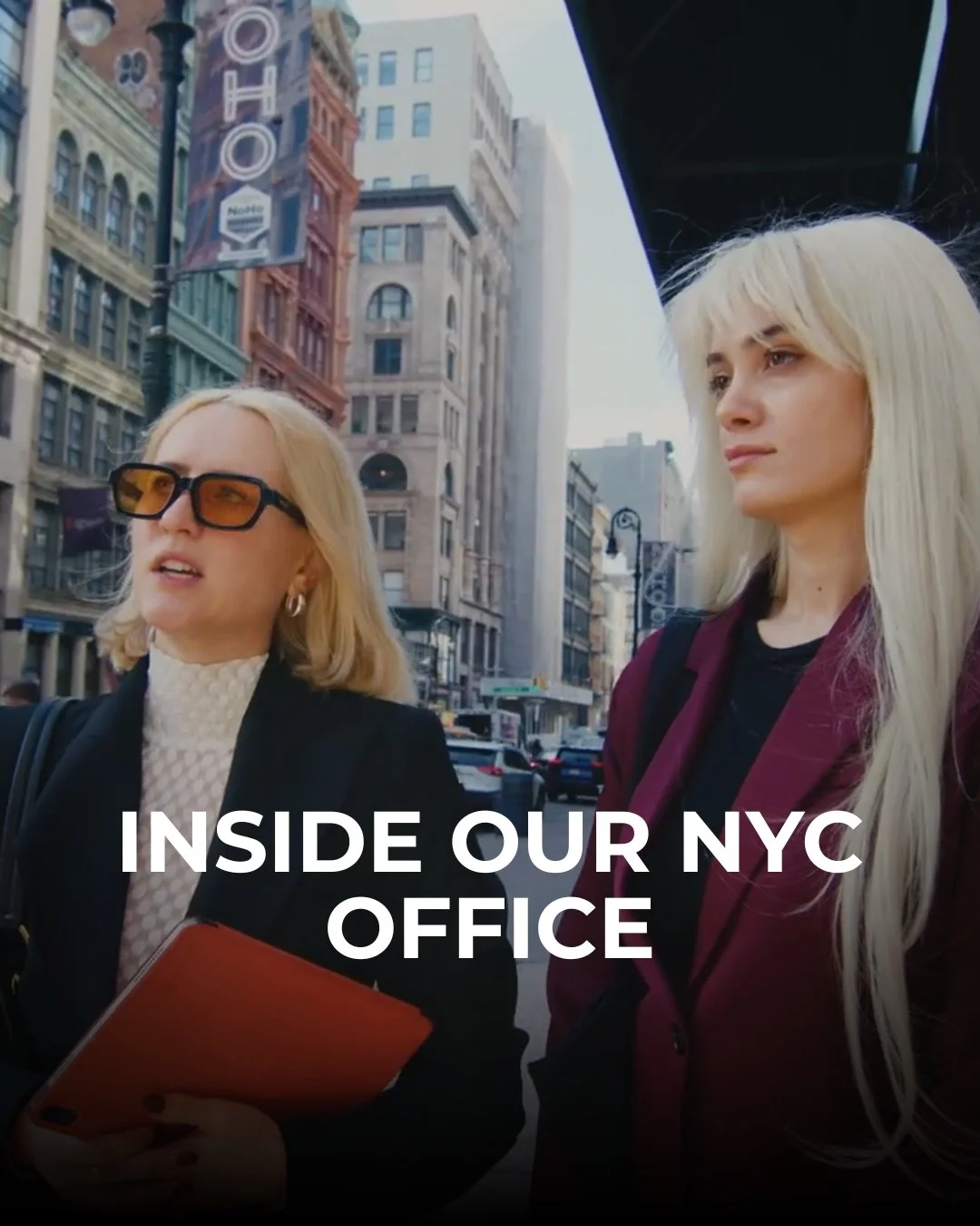 Two women with blonde hair standing on a city street in NYC, surrounded by tall, historic buildings, with cars in the background. One woman is wearing sunglasses and holding a folder, and the other is looking thoughtfully into the distance. The text 