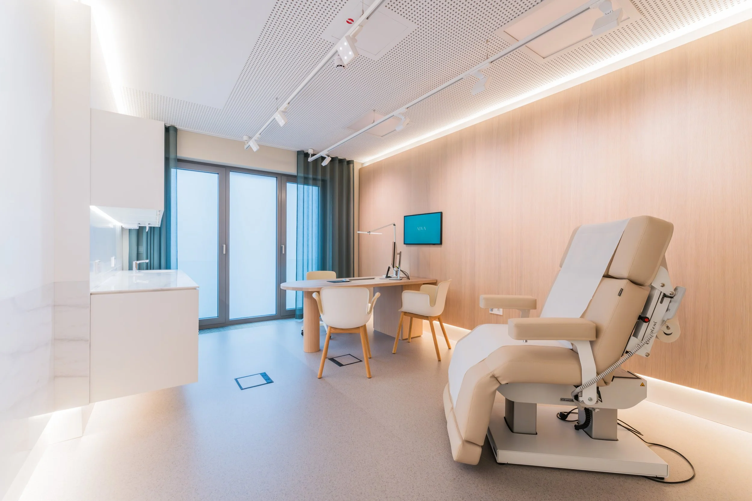 Medical examination room with a reclining examination chair, a desk with computer and chairs, a wall-mounted monitor, and large windows with curtains.