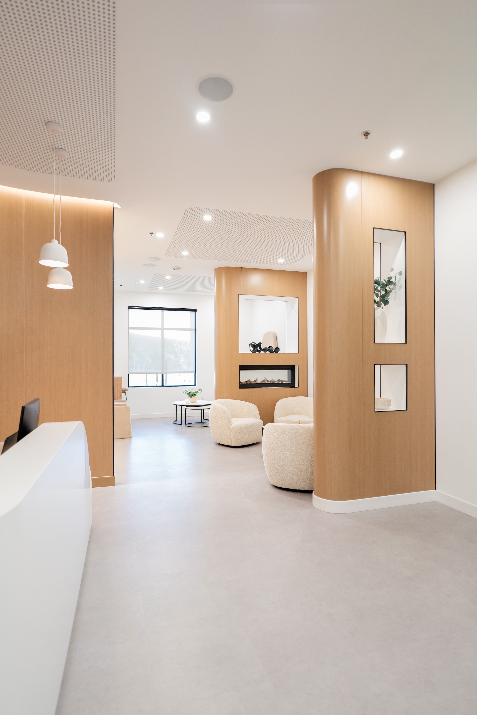 Modern, minimalist interior space with wooden partitions, white seating, a fireplace, potted plant, and large window.