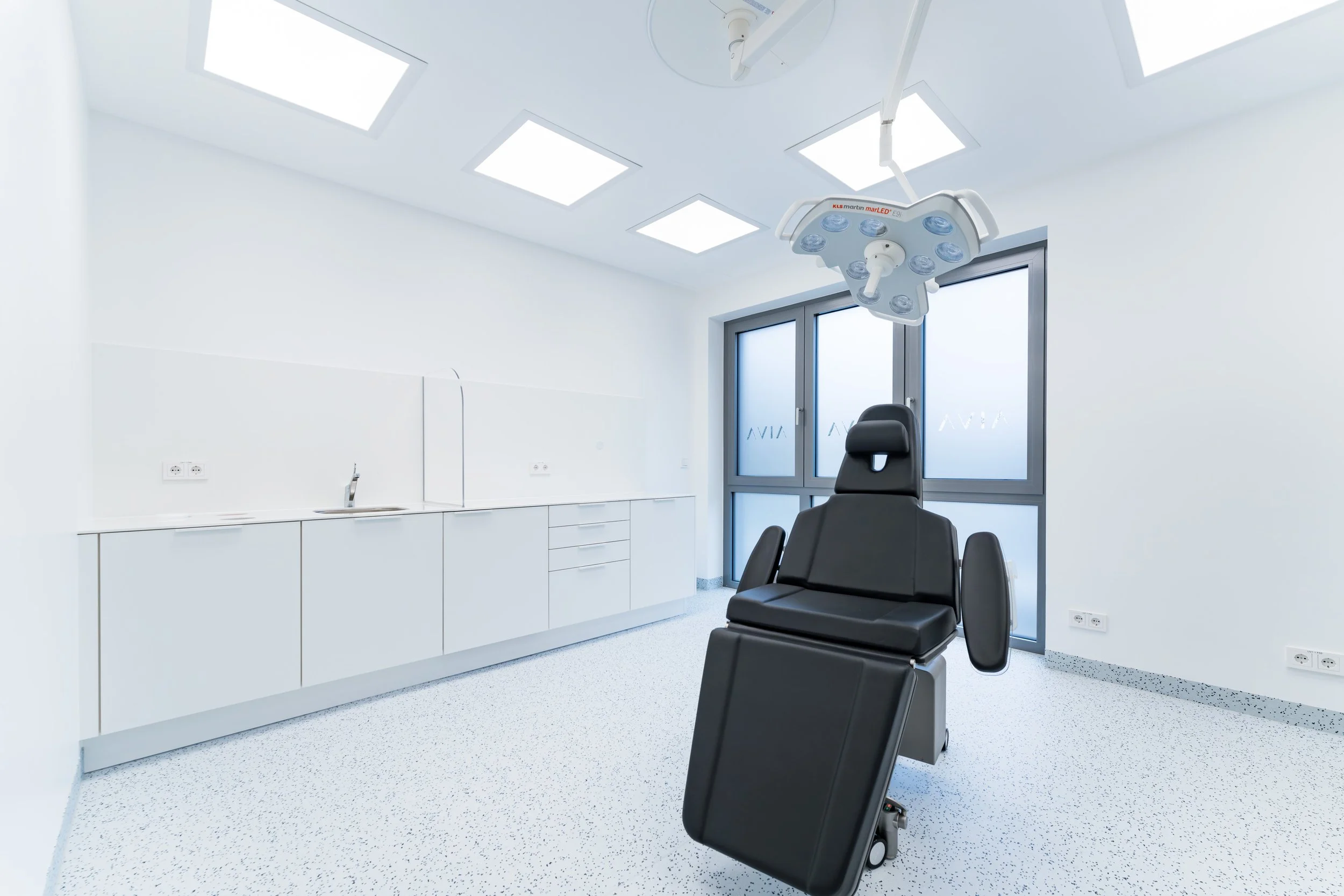 Empty medical examination room with a reclining chair, overhead surgical lights, white cabinetry, a sink, and large windows with frosted glass.