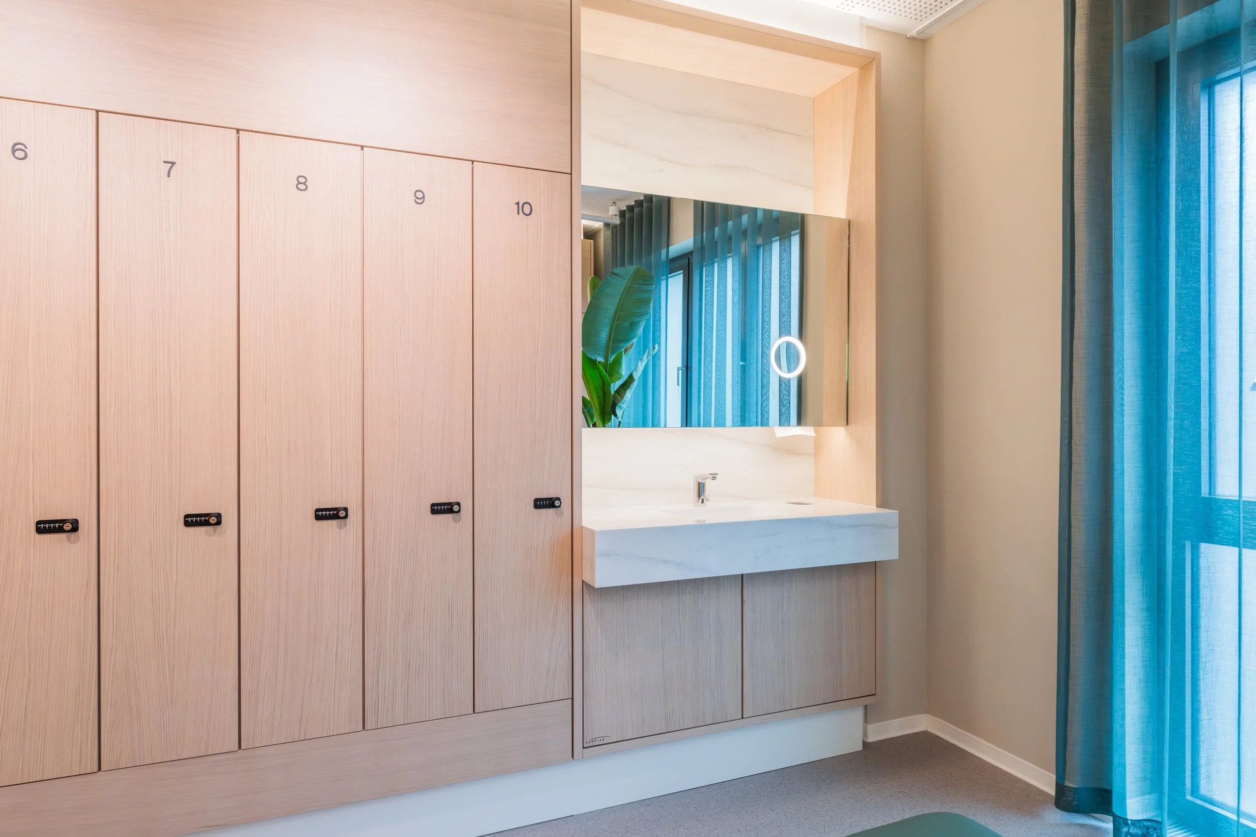 Modern indoor space with wooden lockers numbered 6 to 10 on the left, a white marble sink with a large mirror above, a green plant reflection in the mirror, and a glass door with teal curtain on the right.