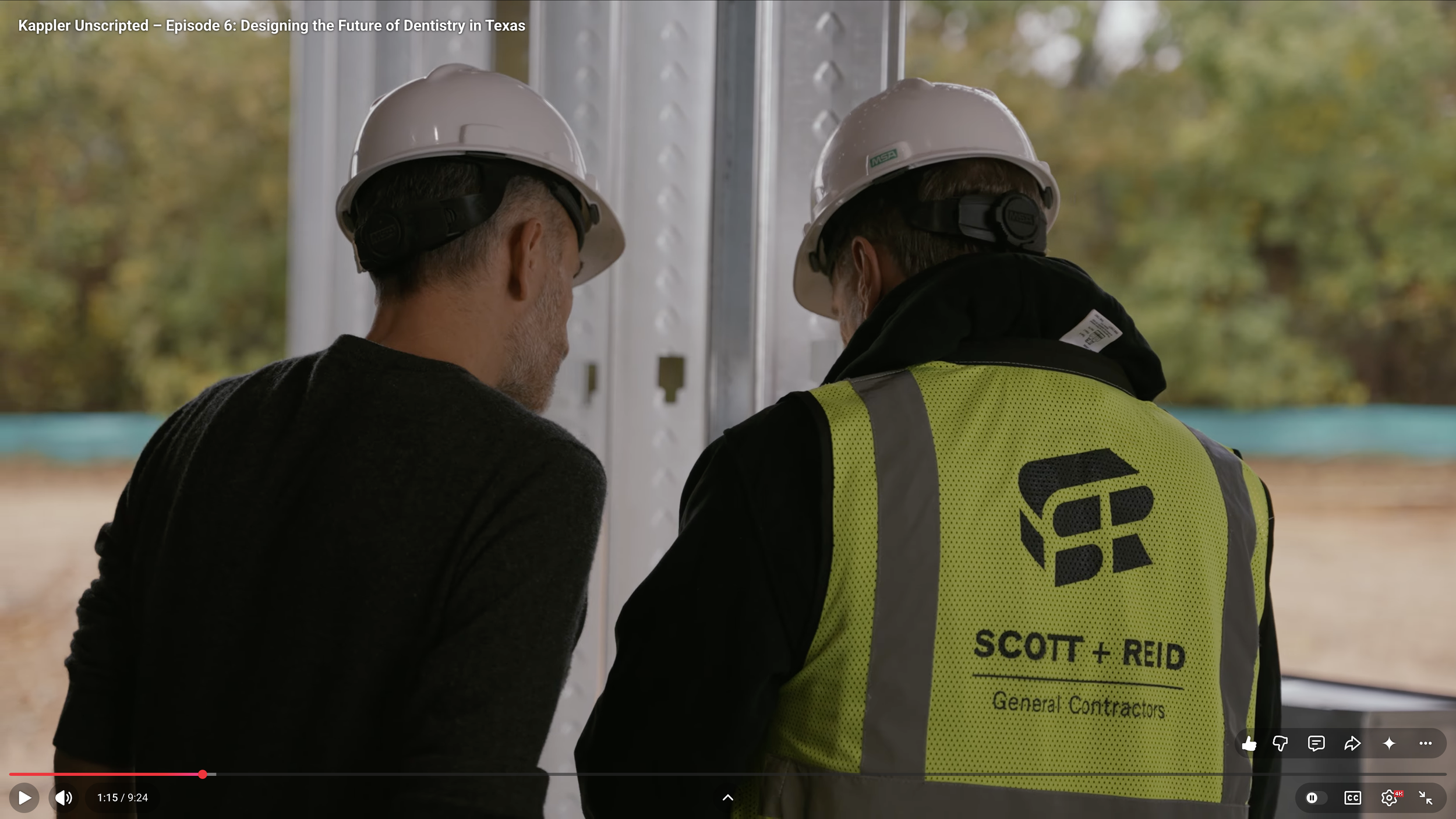 Two construction workers wearing white hard hats and safety gear standing next to a metallic wall, talking outdoors with trees in the background.