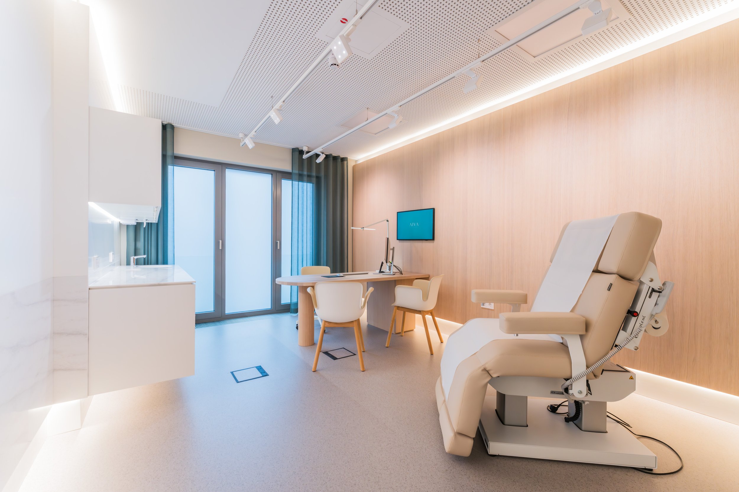 Medical examination room with a hospital bed, a desk with a computer, chairs, and a flat-screen monitor on the wall.