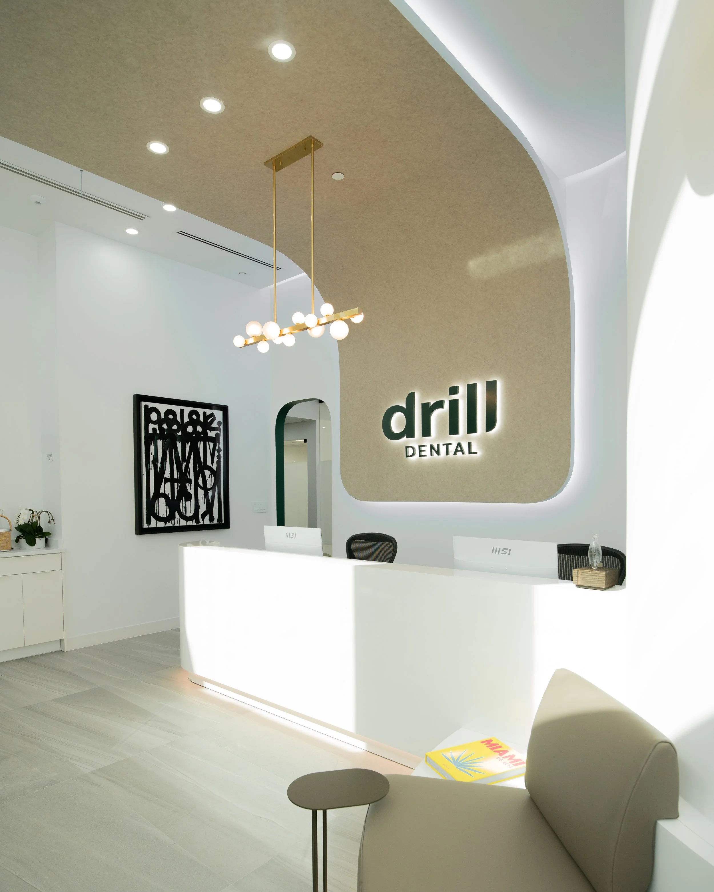 Reception area of a dental office with a white counter, a black chair, a small side table, modern lighting fixtures, and a decorative framed art piece on the wall. The wall features a large illuminated sign that reads 'drill DENTAL' with a gold and b