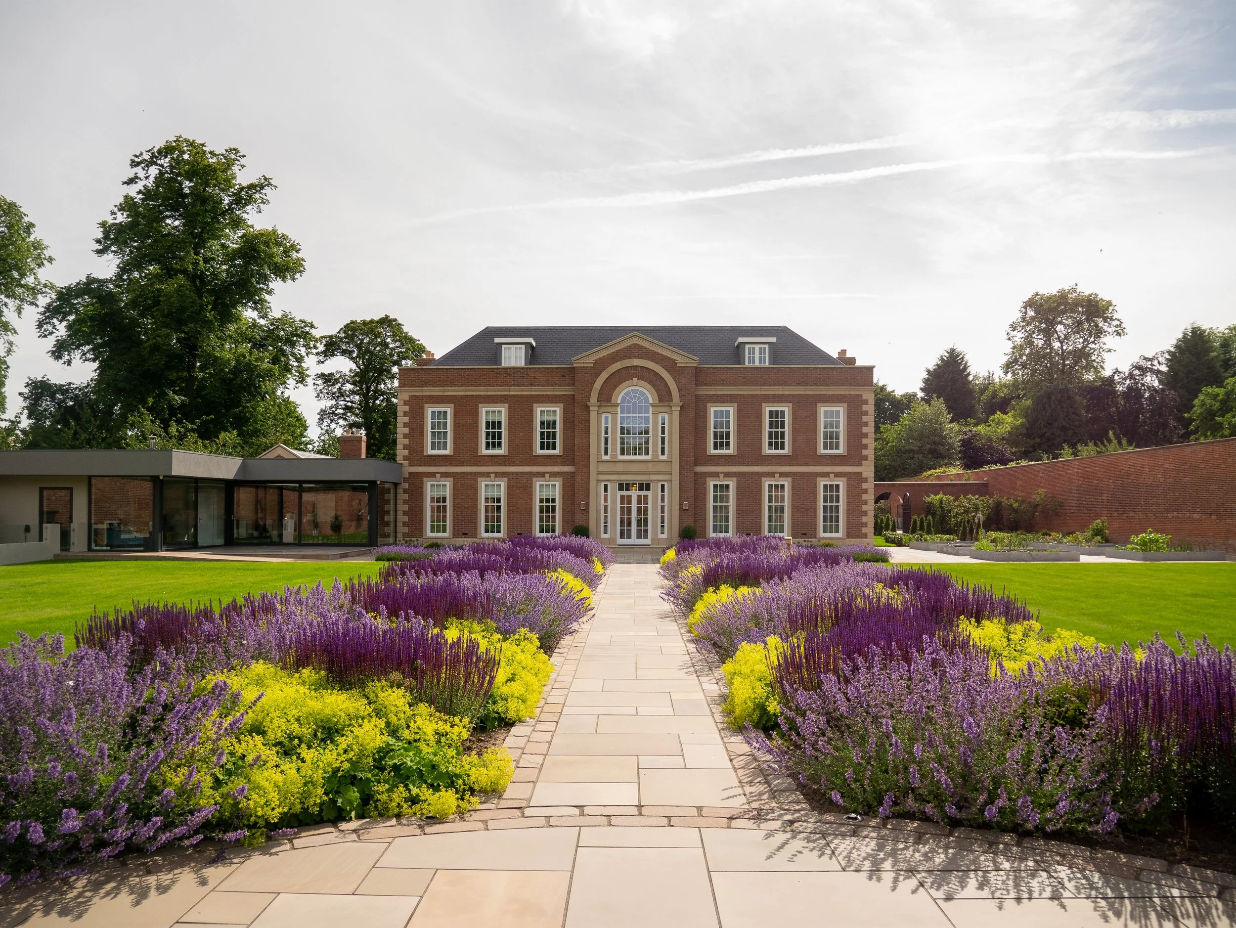 Guy Taylor Associates — A mansion with red brick walls and large windows, surrounded by a garden with purple and yellow flowers along a stone pathway.