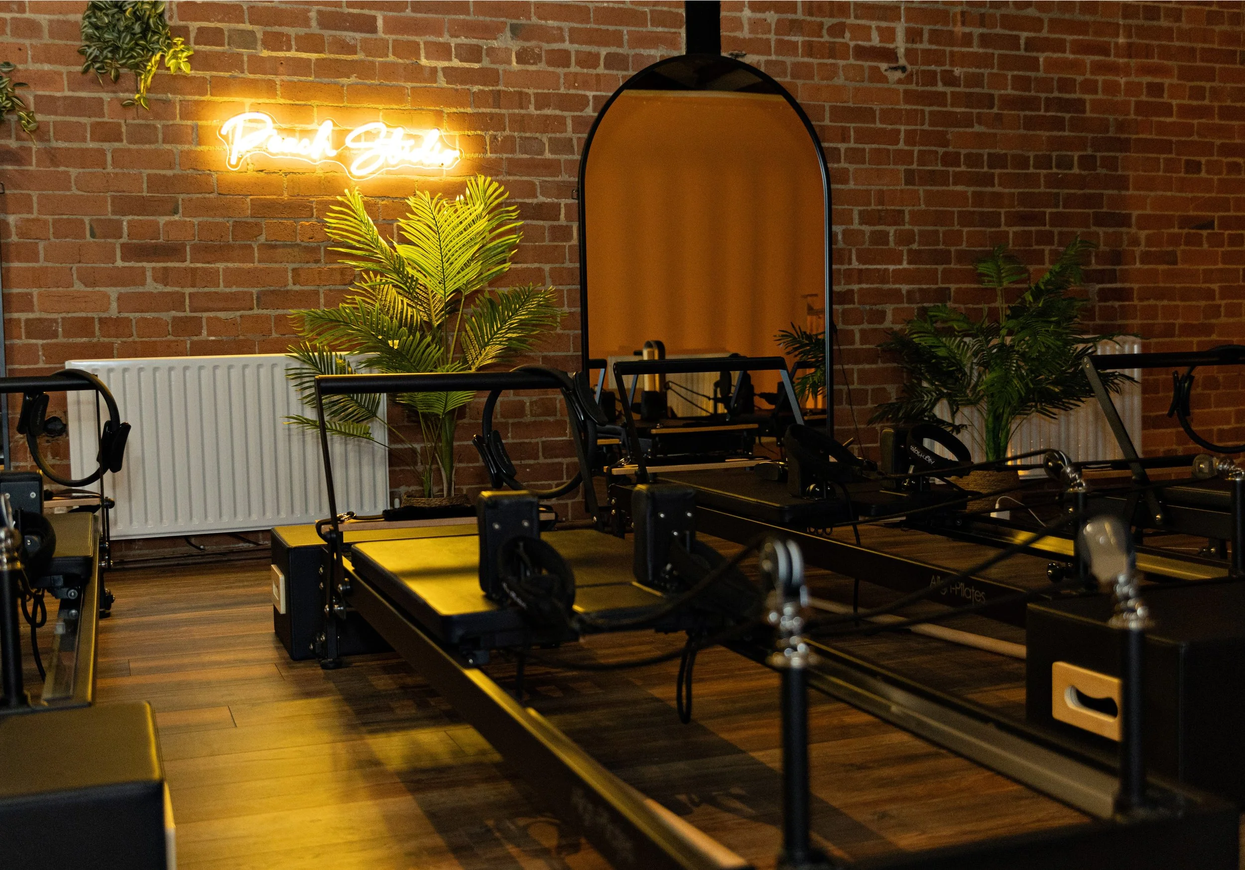 Fitness studio with reformer Pilates equipment, brick wall, neon sign reading 'Pure Studio', and green plants.