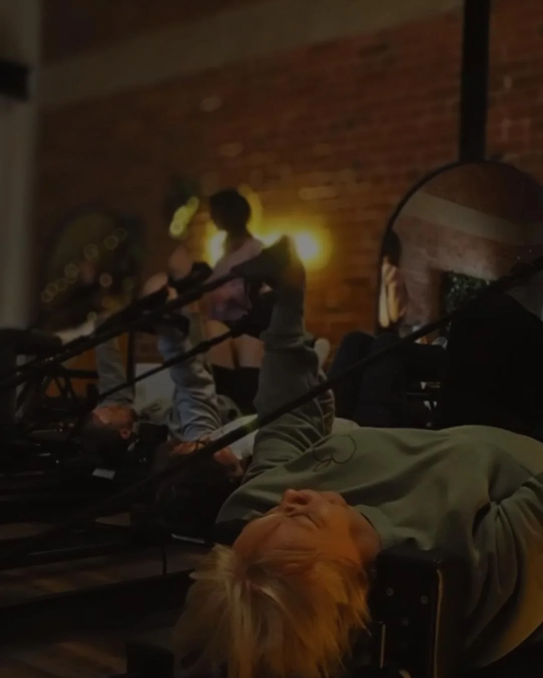 People relaxing on a bed in a dimly lit room with a brick wall, some people are sitting and talking, one person with blond hair is lying down.
