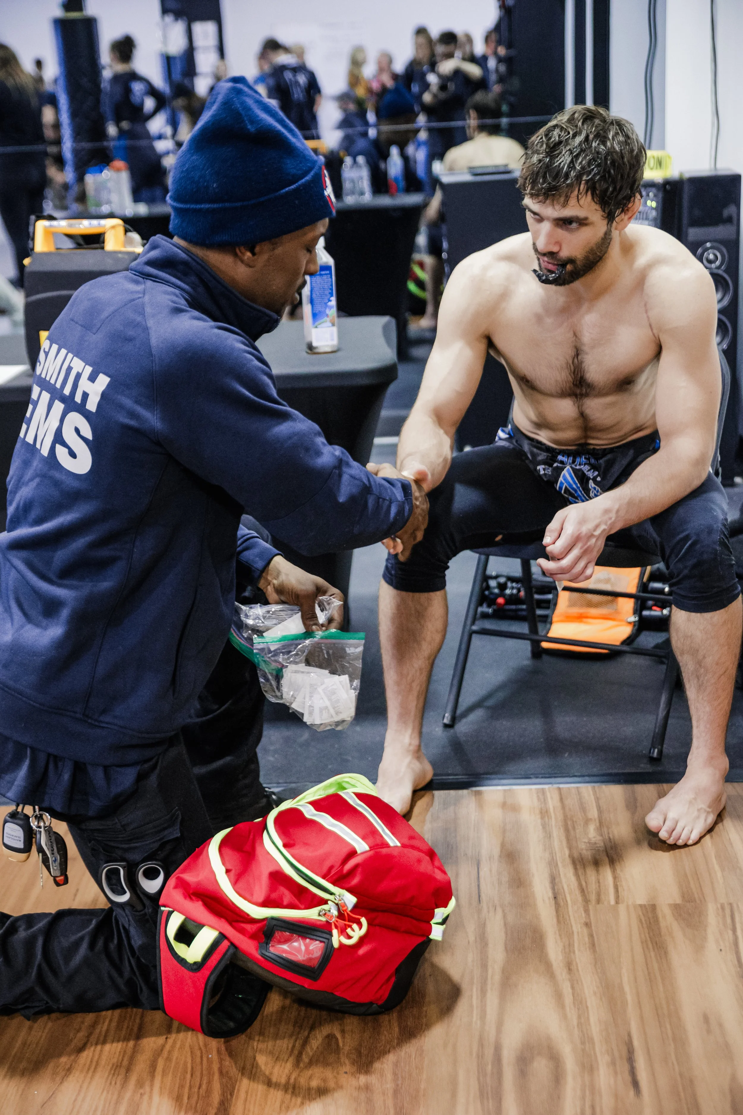 A jiu jitsu competitor sitting on a chair, getting treated by a Smith EMS EMT, inside a busy gym or training area.