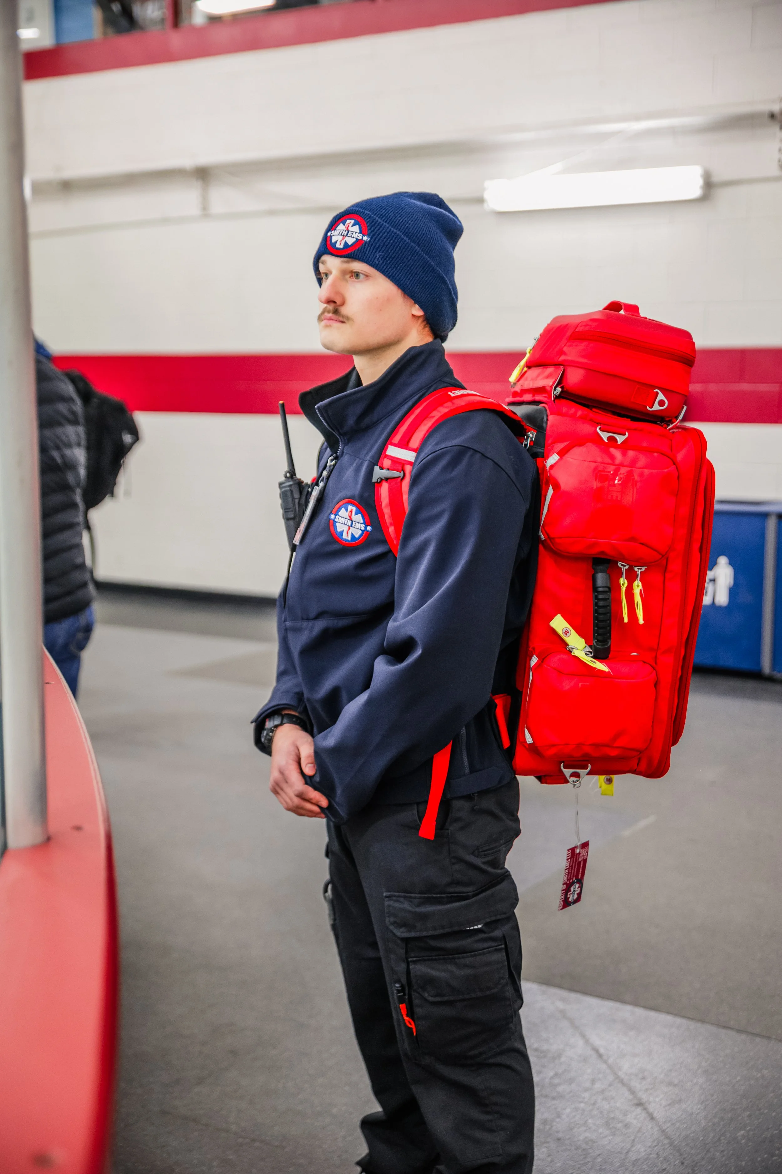 A man wearing a blue beanie and a dark blue jacket with an emergency responder emblem, standing indoors with a red rescue backpack.