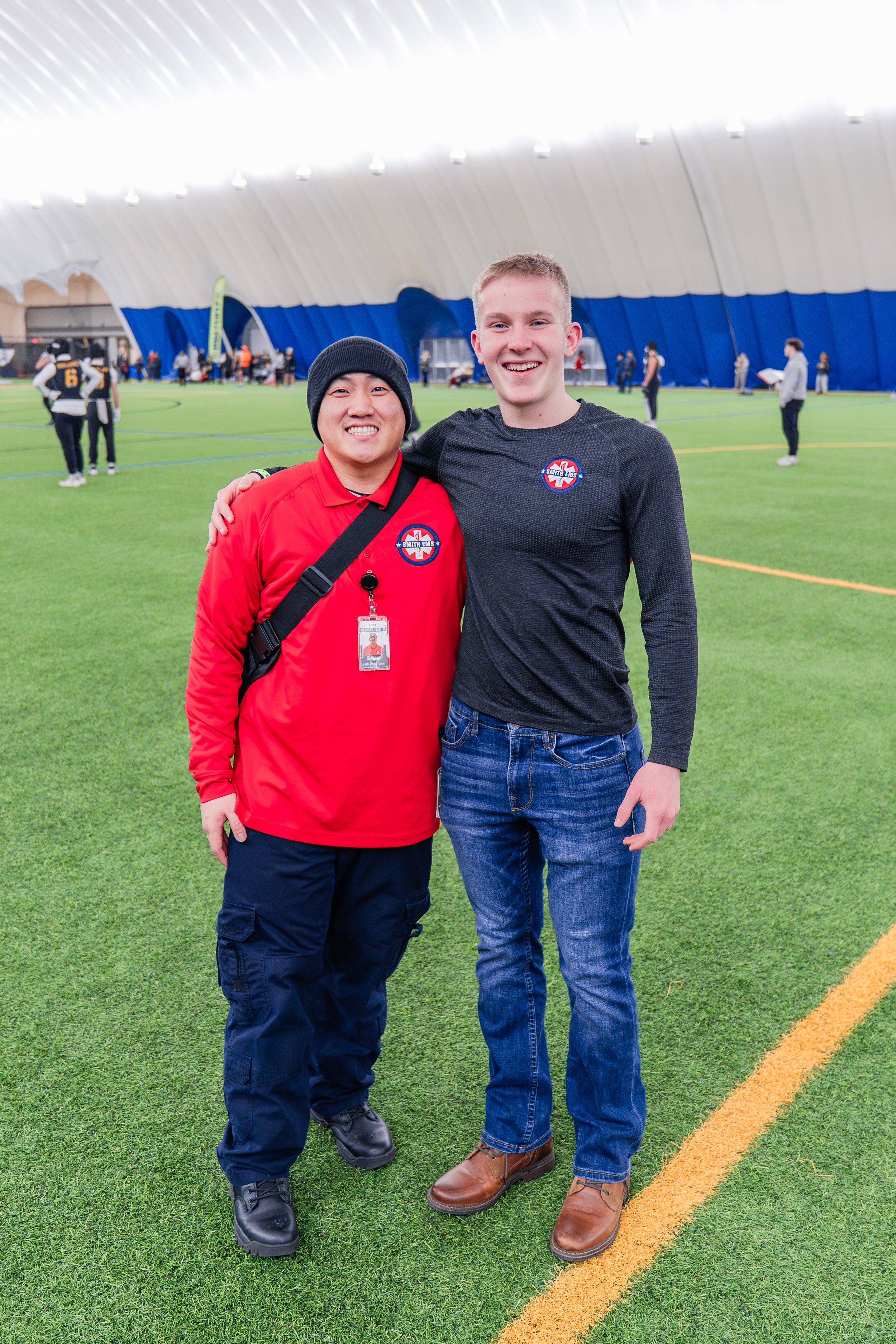 Two men standing close together on indoor artificial turf, smiling at the camera, with a large curved ceiling and other people in the background.