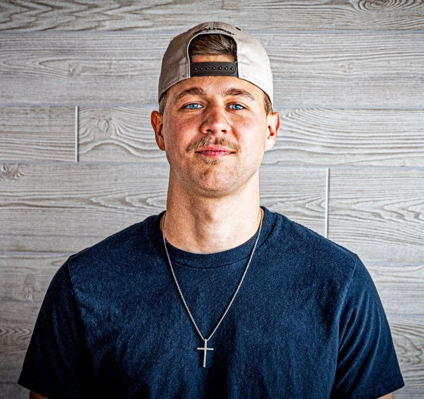 Young man with blue eyes wearing a backward baseball cap, a black t-shirt, and a necklace with a cross, standing in front of a light wood-paneled wall.