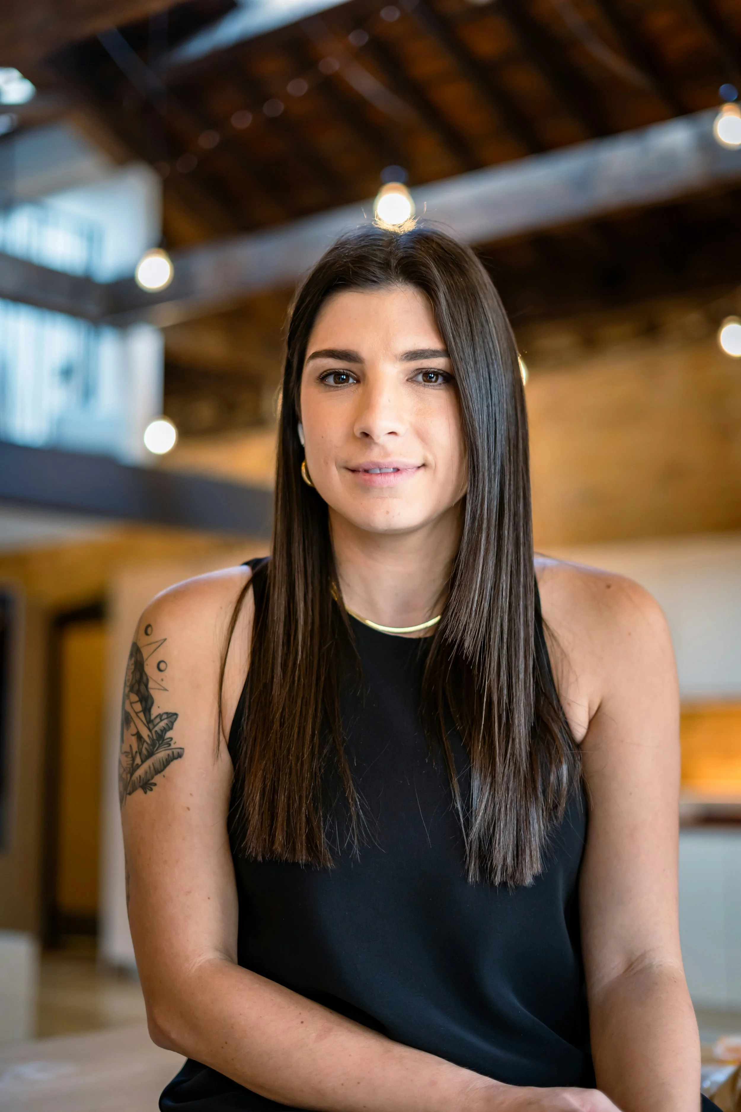 A young woman with straight brown hair and a tattoo on her upper arm, wearing a black sleeveless top, sitting indoors with blurred lights and wooden ceiling in the background.