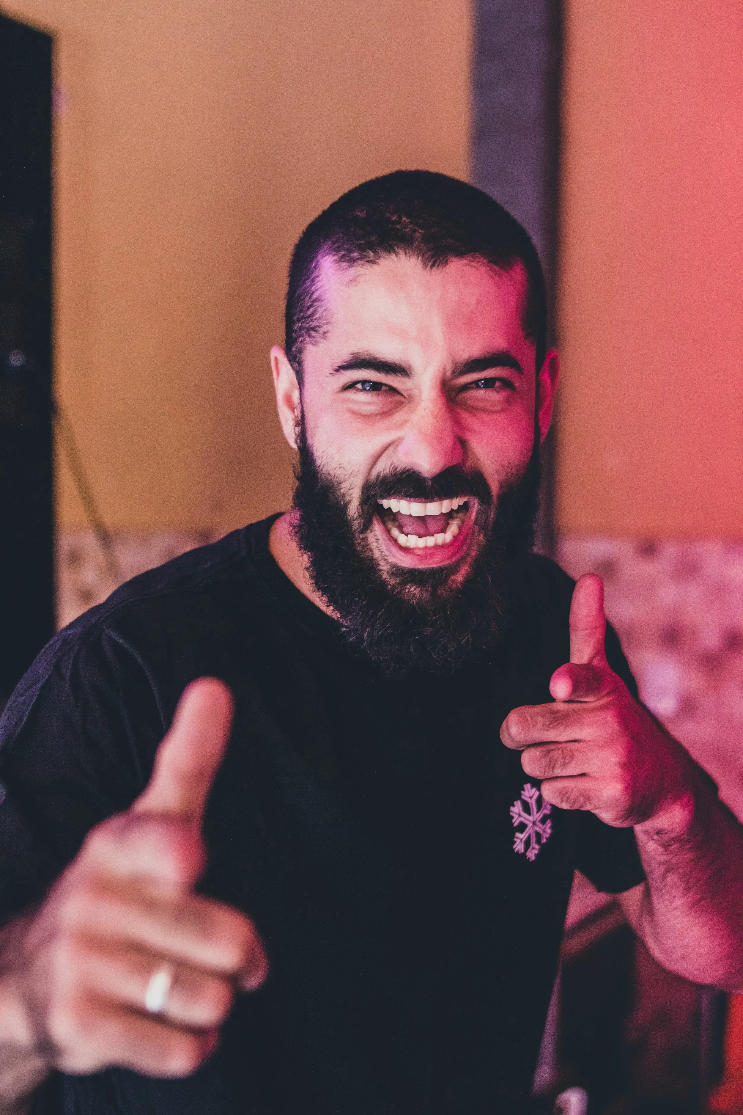 A man with a beard making a thumbs-up gesture, smiling and pointing with his index finger, in a lively indoor setting.
