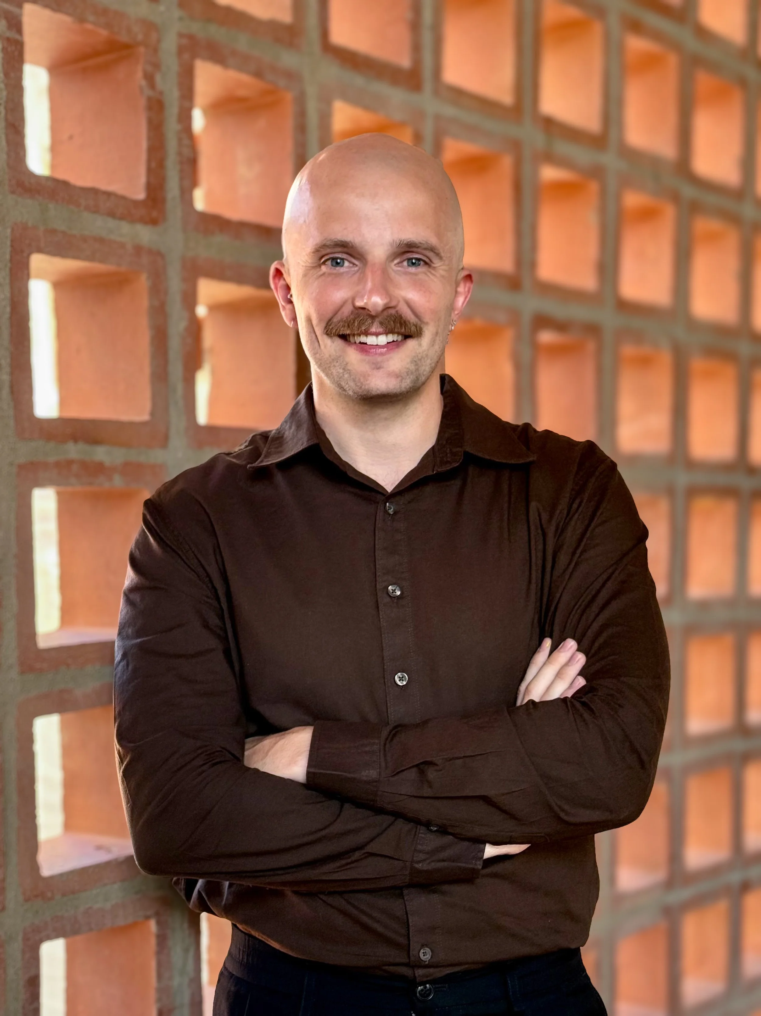 Man with a bald head, mustache, and jewelry in front of a brick wall with square openings, crossing his arms and smiling.