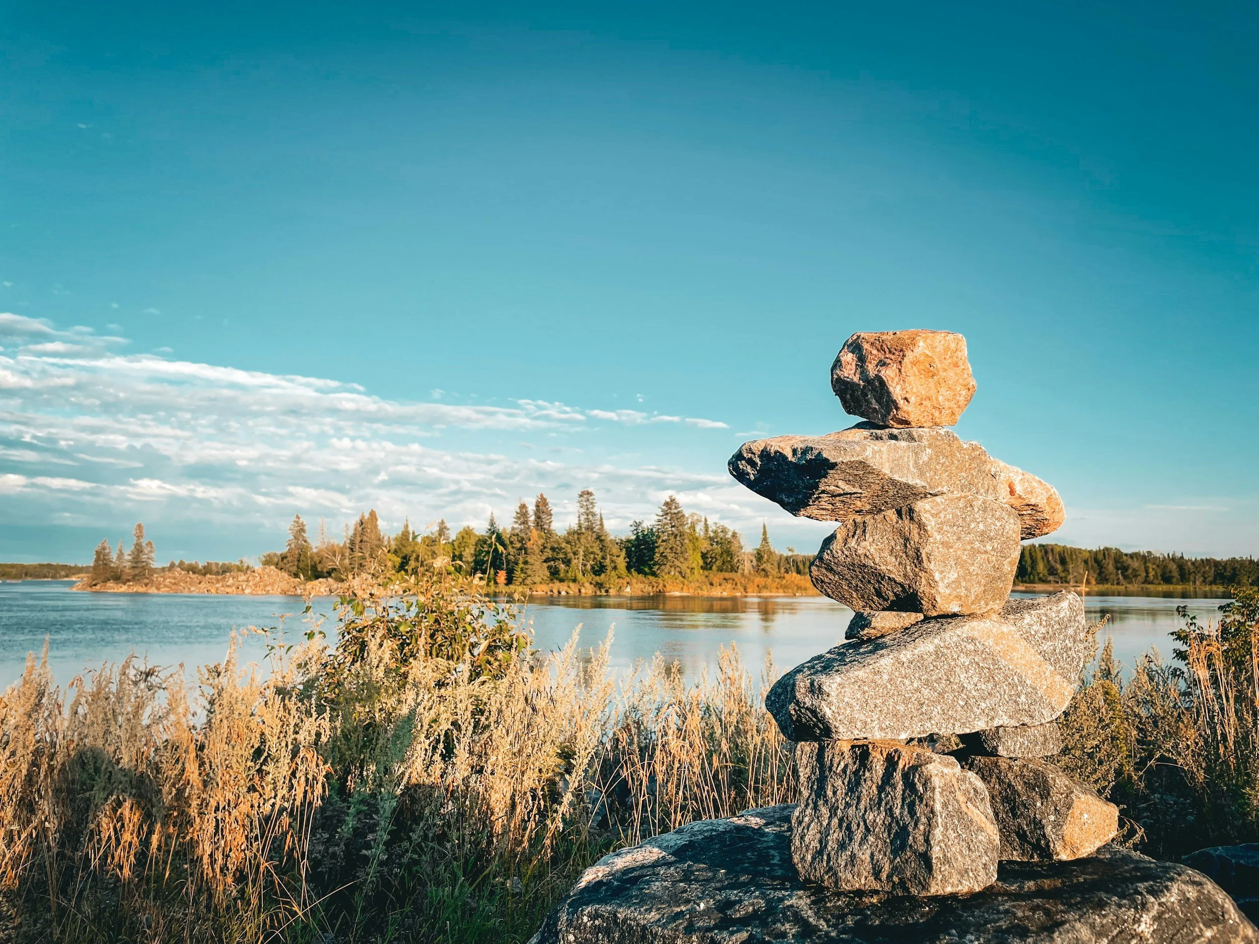 Stacked rocks in the foreground near a lake with trees and a partly cloudy sky in the background.