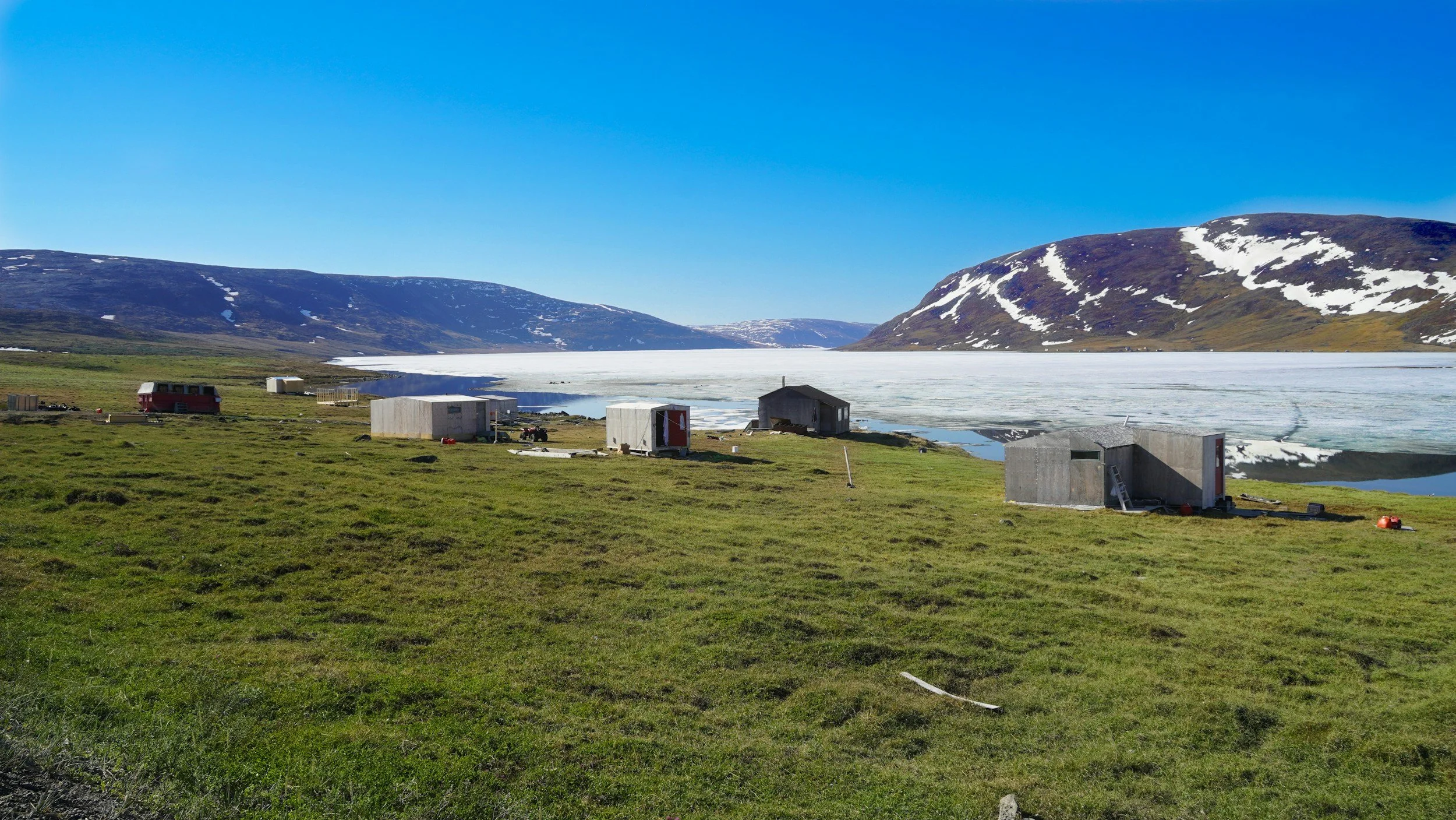 A landscape with green grass, several small buildings, a frozen body of water, and snow-capped mountains under a clear blue sky.