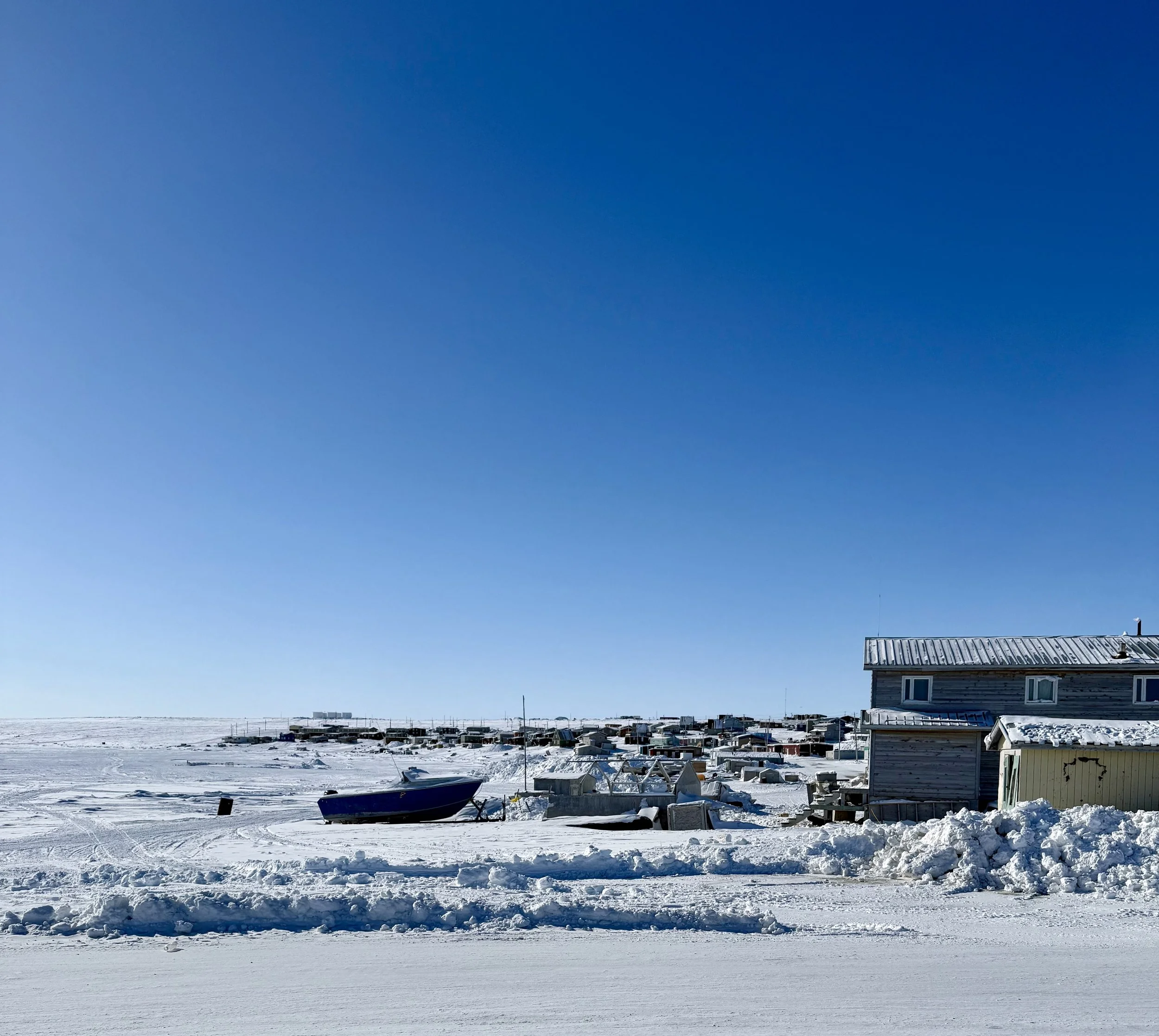 Snow-covered landscape with a house and boat, under a clear blue sky, with distant buildings on the horizon.