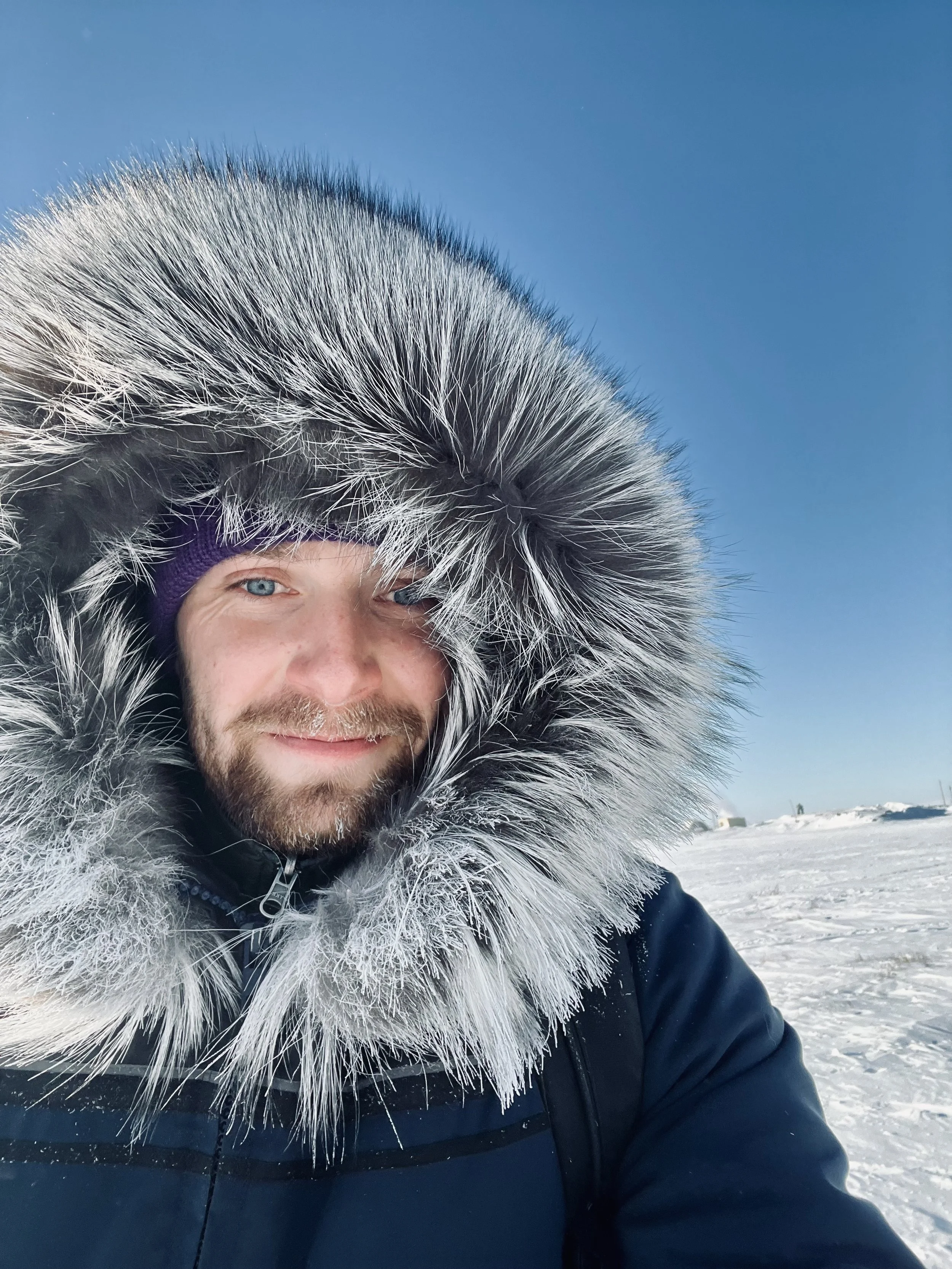 Man in a snowy landscape wearing a parka with a fur-lined hood, smiling at the camera.