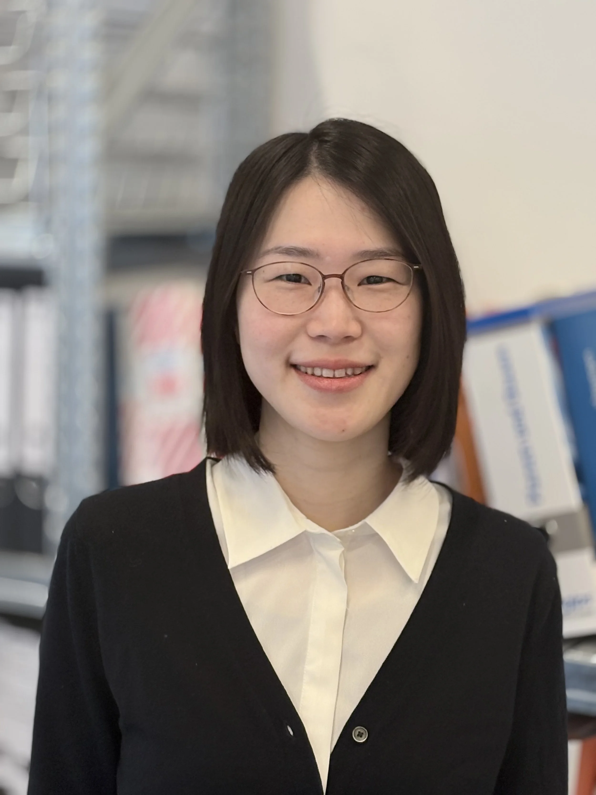 Eine lachende junge Frau mit schwarzen Haaren, tragt einen grauen Pullover und steht in einem Raum mit Büchern im Hintergrund.