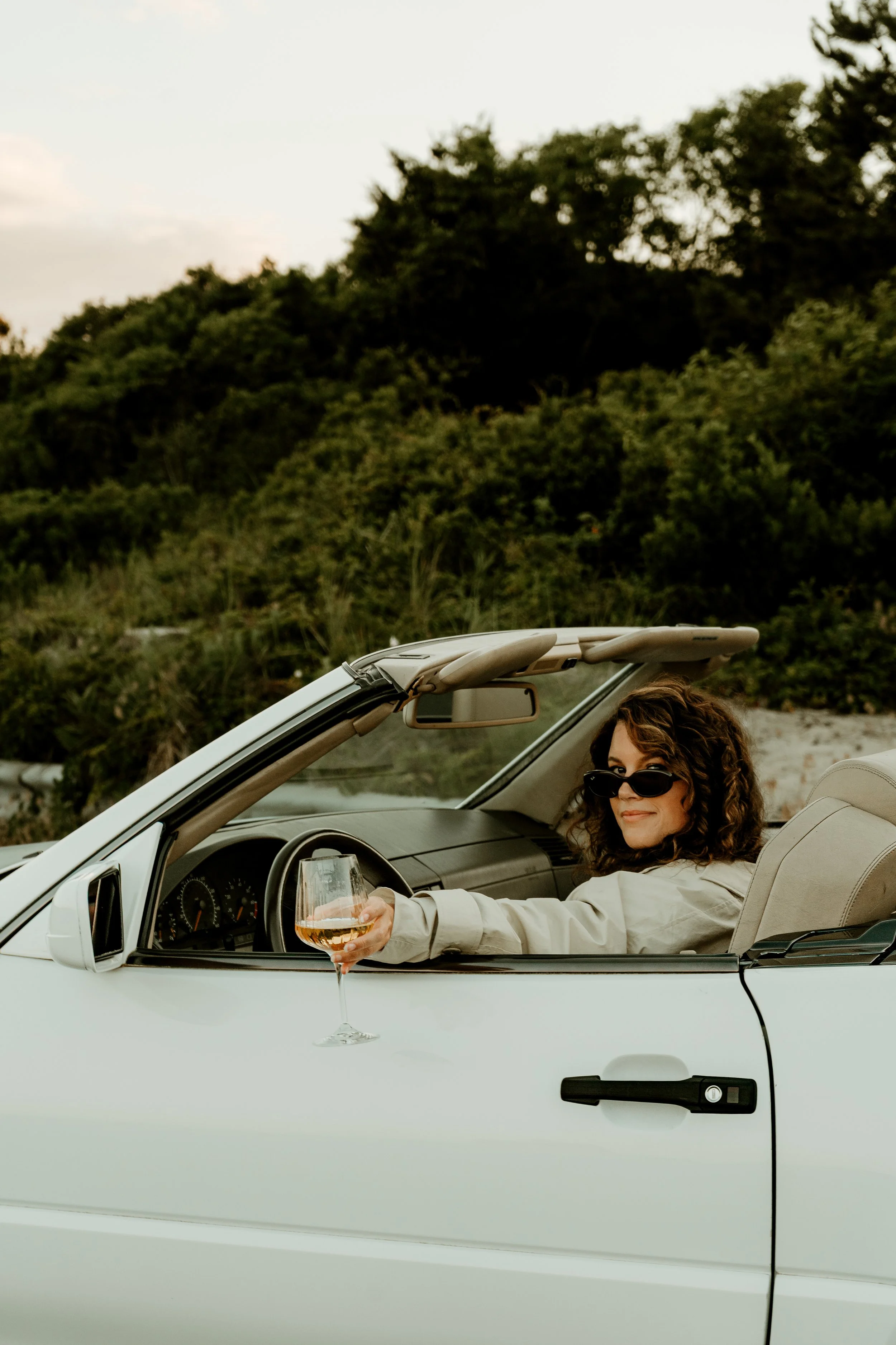 A woman with curly hair and sunglasses sitting in a white convertible car, holding a glass of rosé wine, parked outdoors with green hills and trees in the background during daytime.