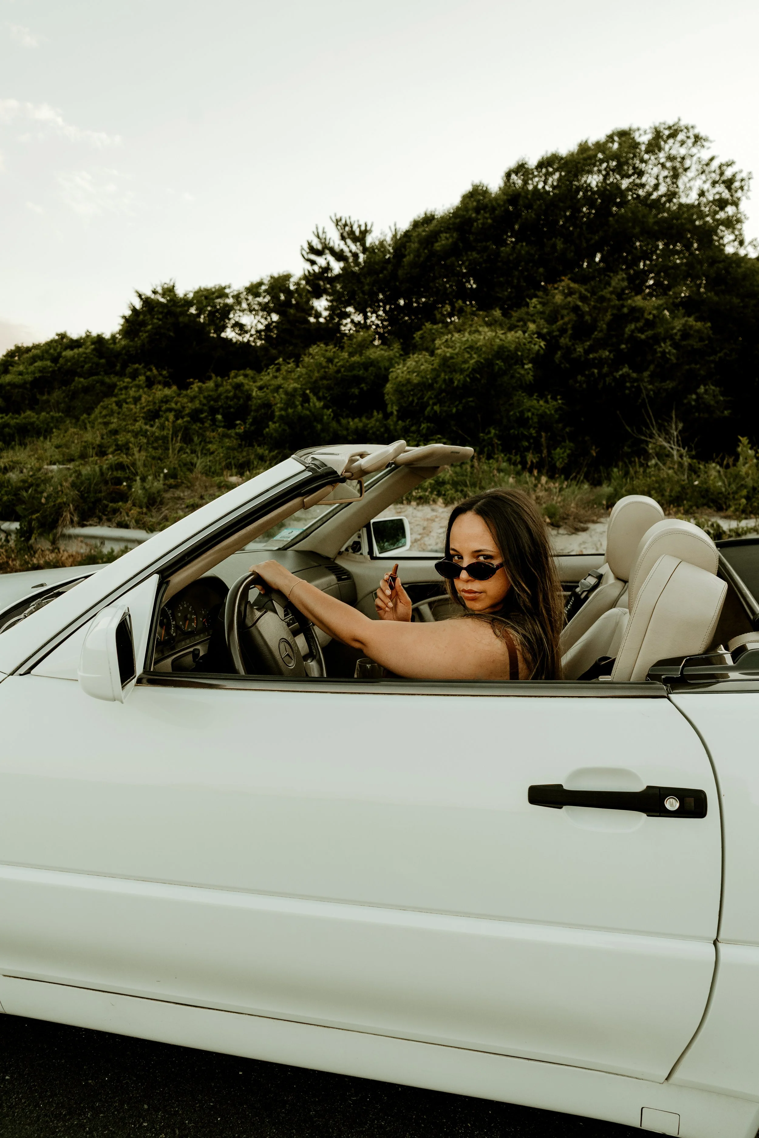 A woman with dark hair and sunglasses sitting in the driver's seat of a white convertible car, holding a small object in her right hand, with greenery and a clear sky in the background.