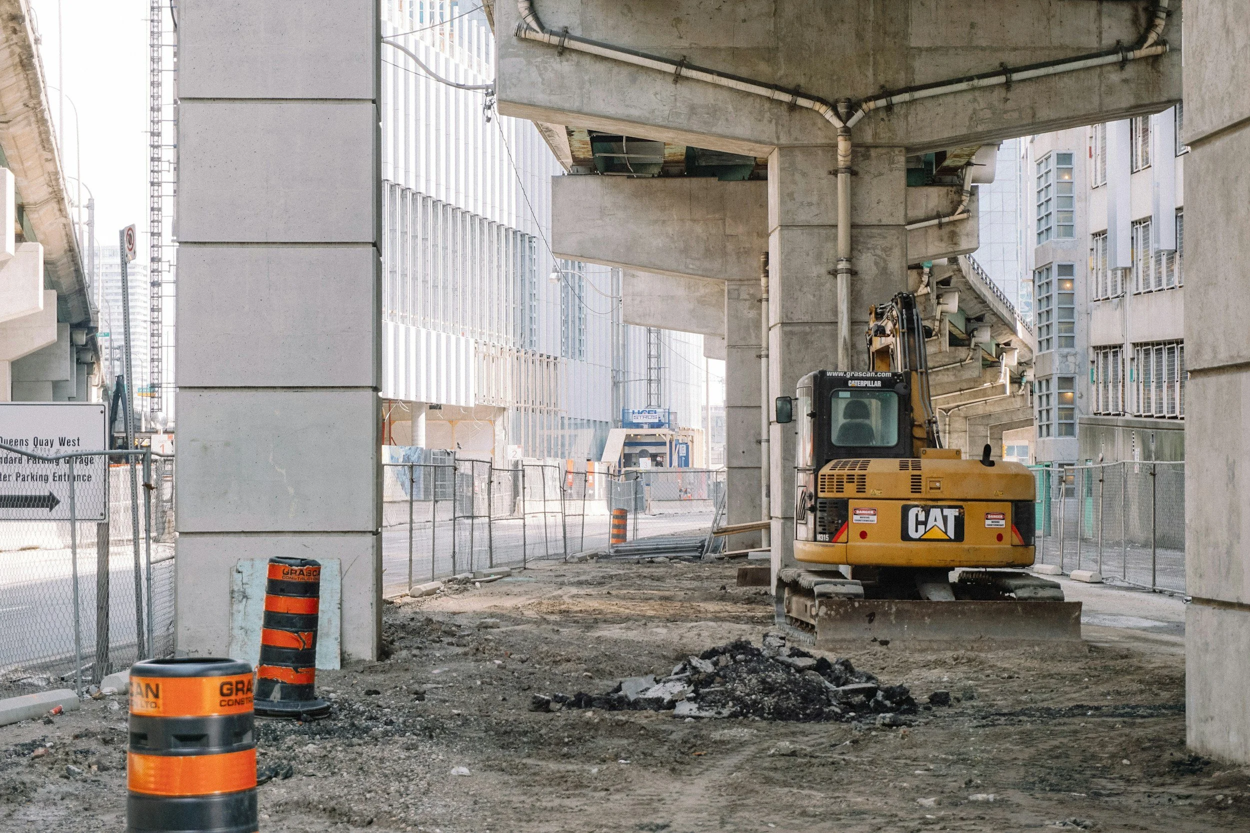 Construction site under a bridge with a yellow excavator parked on dirt, surrounded by orange cones and metal fencing.