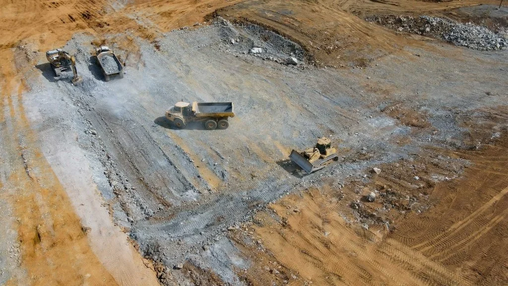 Construction site with heavy machinery, including an excavator, dump trucks, and bulldozer working on earthmoving and excavation.