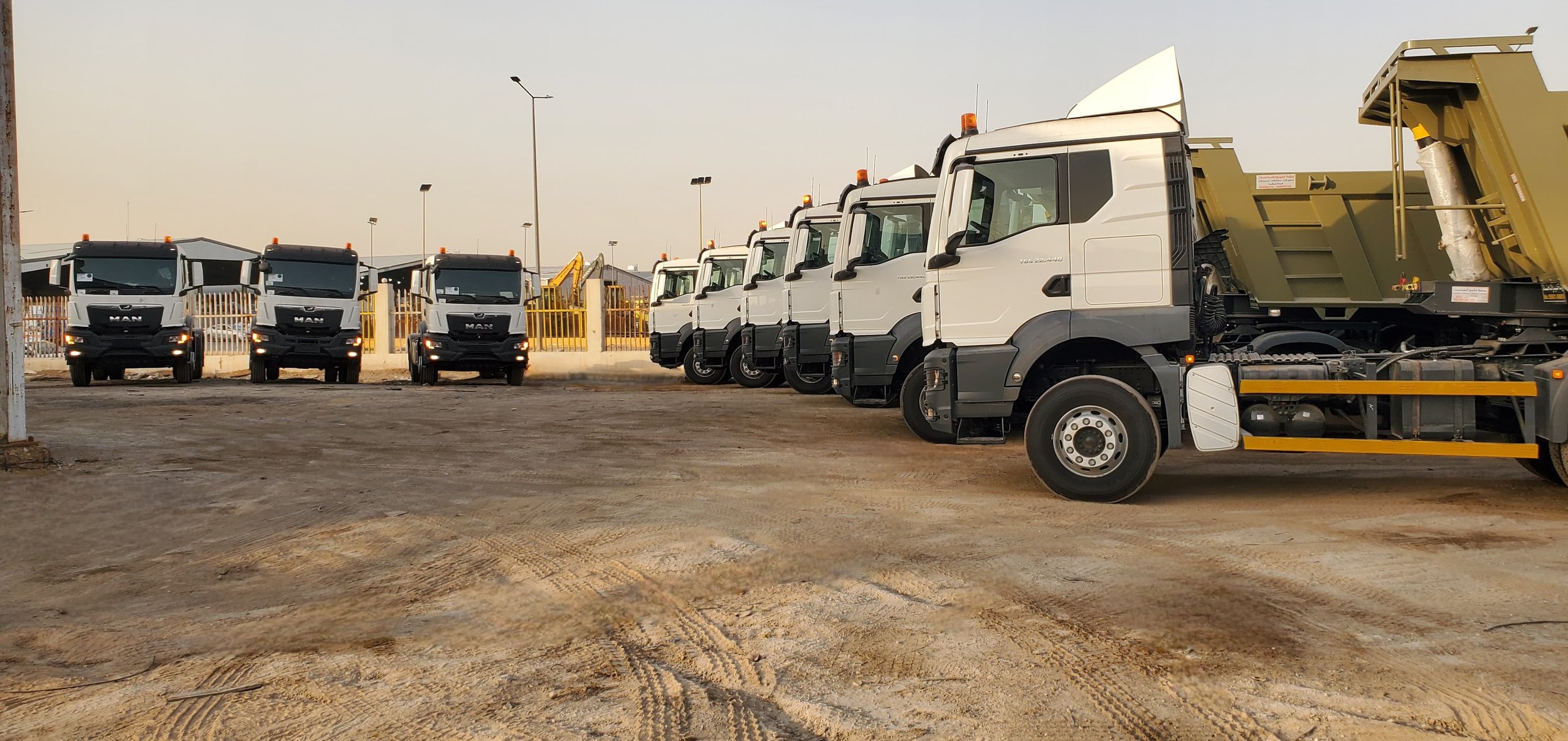 Row of white dump trucks parked on a dirt lot with construction equipment in the background.