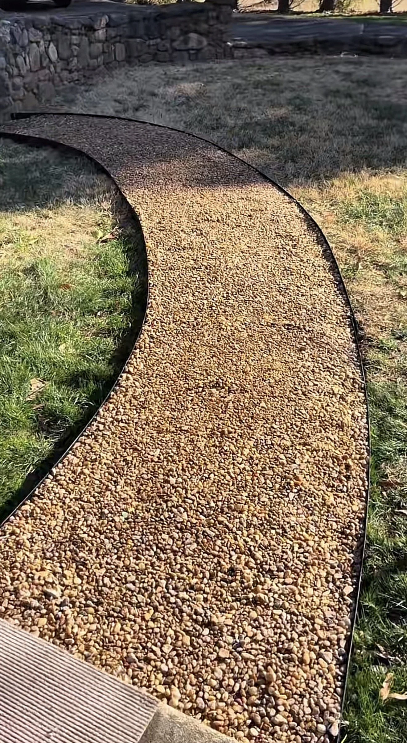 A curved pathway made of small beige and brown gravel, bordered by a black edge, surrounded by grass and a stone wall in the background.