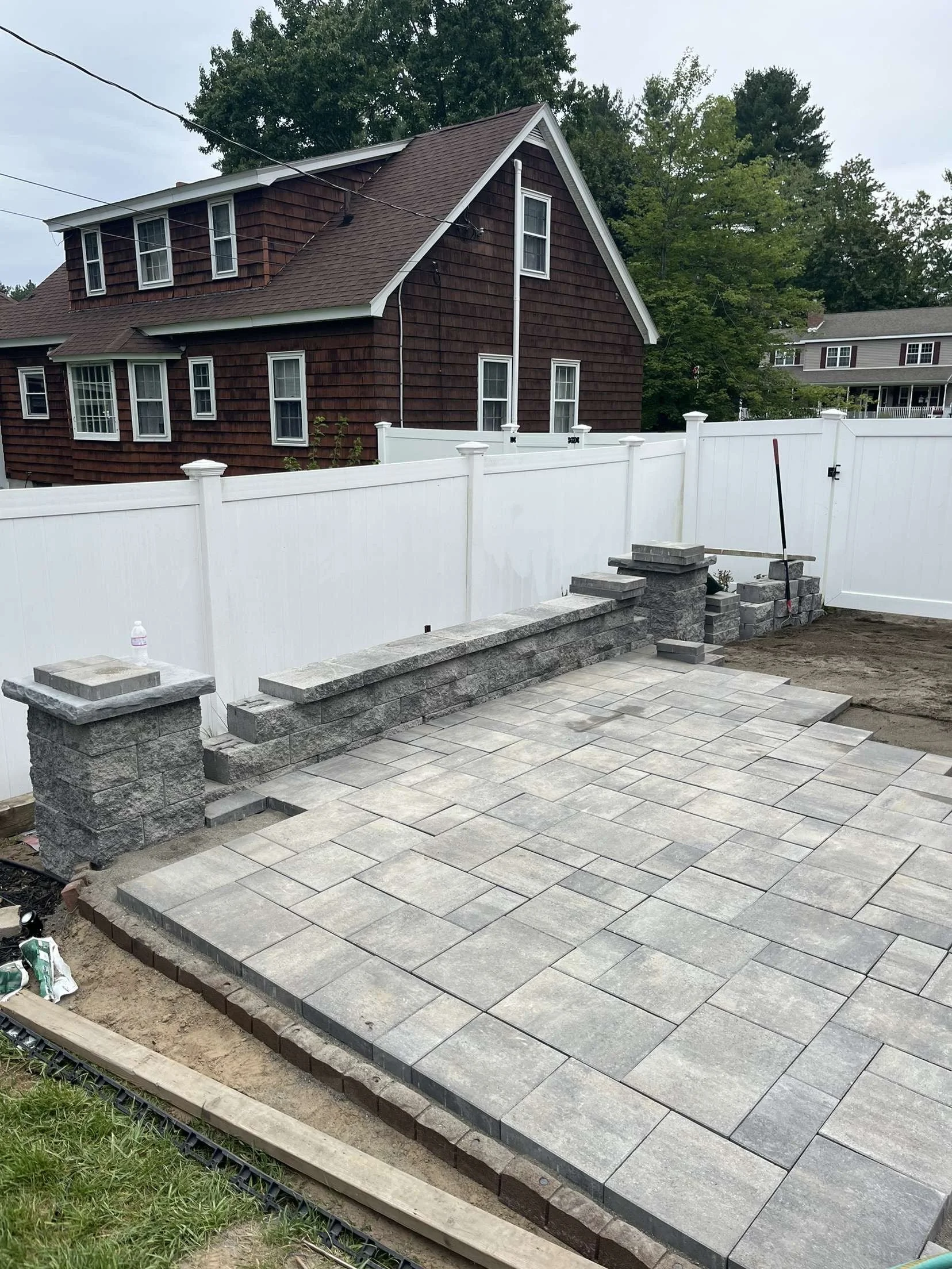 Partially completed outdoor patio with gray stone tiles and a brick wall under construction, surrounded by a white privacy fence.