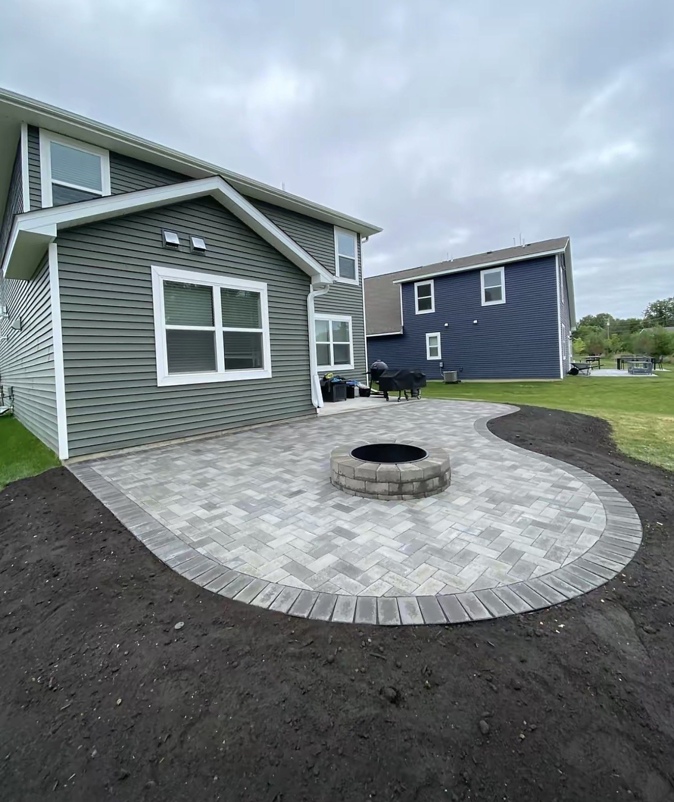 Newly constructed backyard patio with paver stones, a fire pit, and a grill, surrounded by a lawn with some patches of soil, in a residential area with two houses and a cloudy sky.