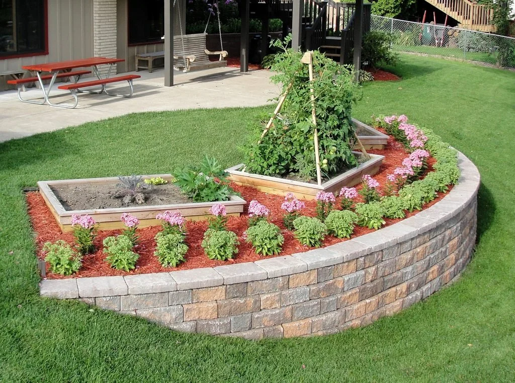 A landscaped backyard with a raised garden bed filled with green plants, surrounded by pink flowering plants, with a brick retaining wall, a green lawn, a patio with a picnic table, and a wooden swing.